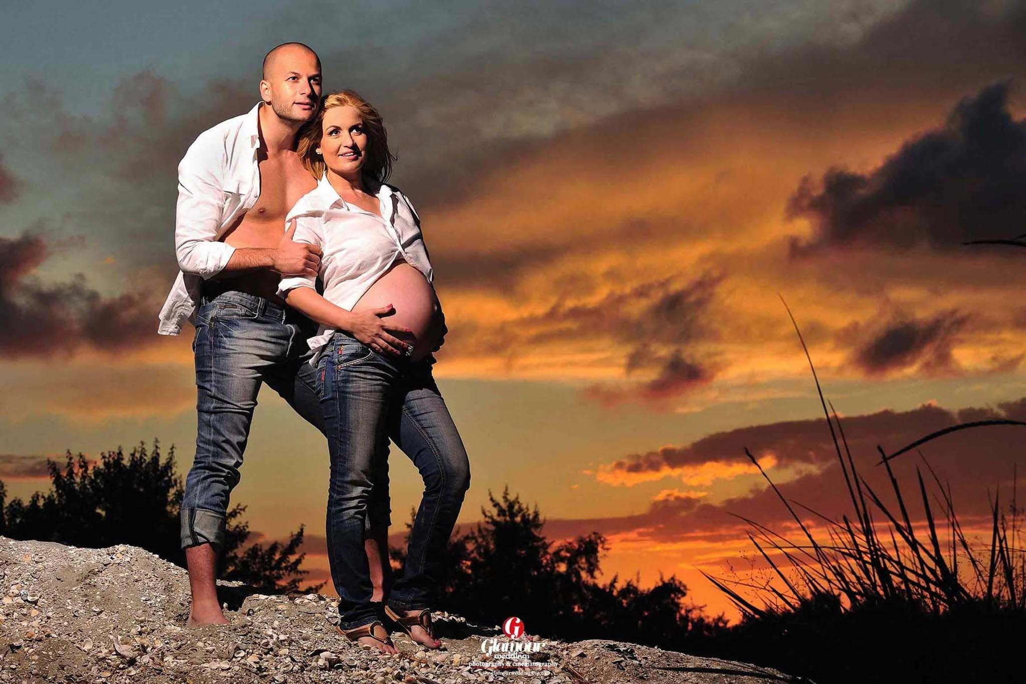 Outdoor maternity portrait of an expectant couple in casual styling against a dramatic sunset sky.