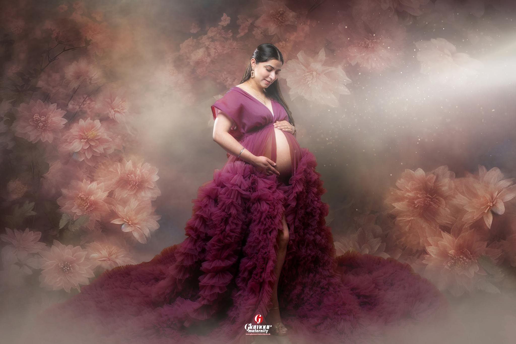 Fine-art maternity portrait in a burgundy ruffled gown surrounded by soft flowers, mist and warm romantic tones.