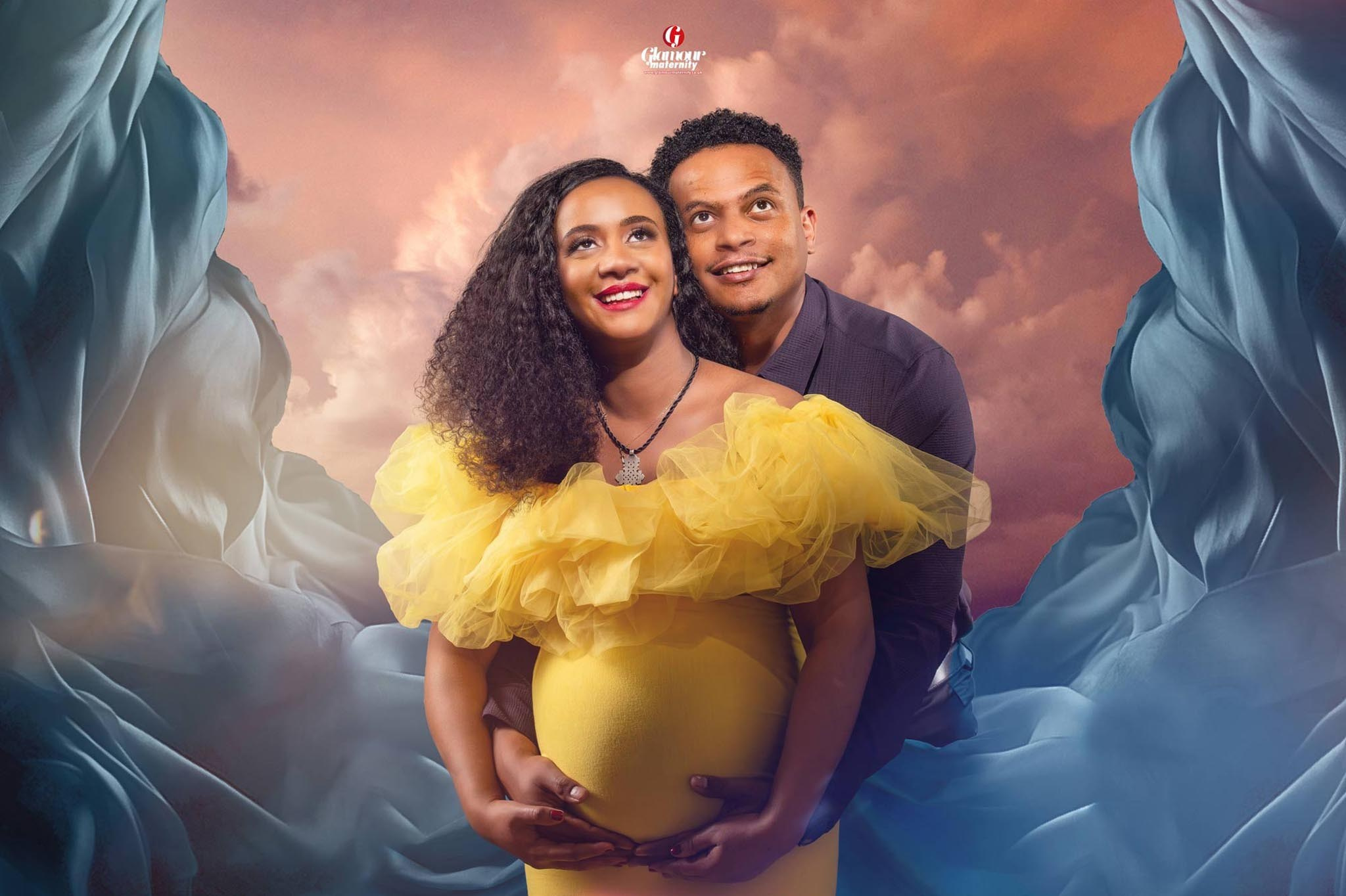 Romantic maternity portrait of an expectant couple, with the woman wearing a yellow ruffled gown, flowing blue fabric and a dreamy sky background.