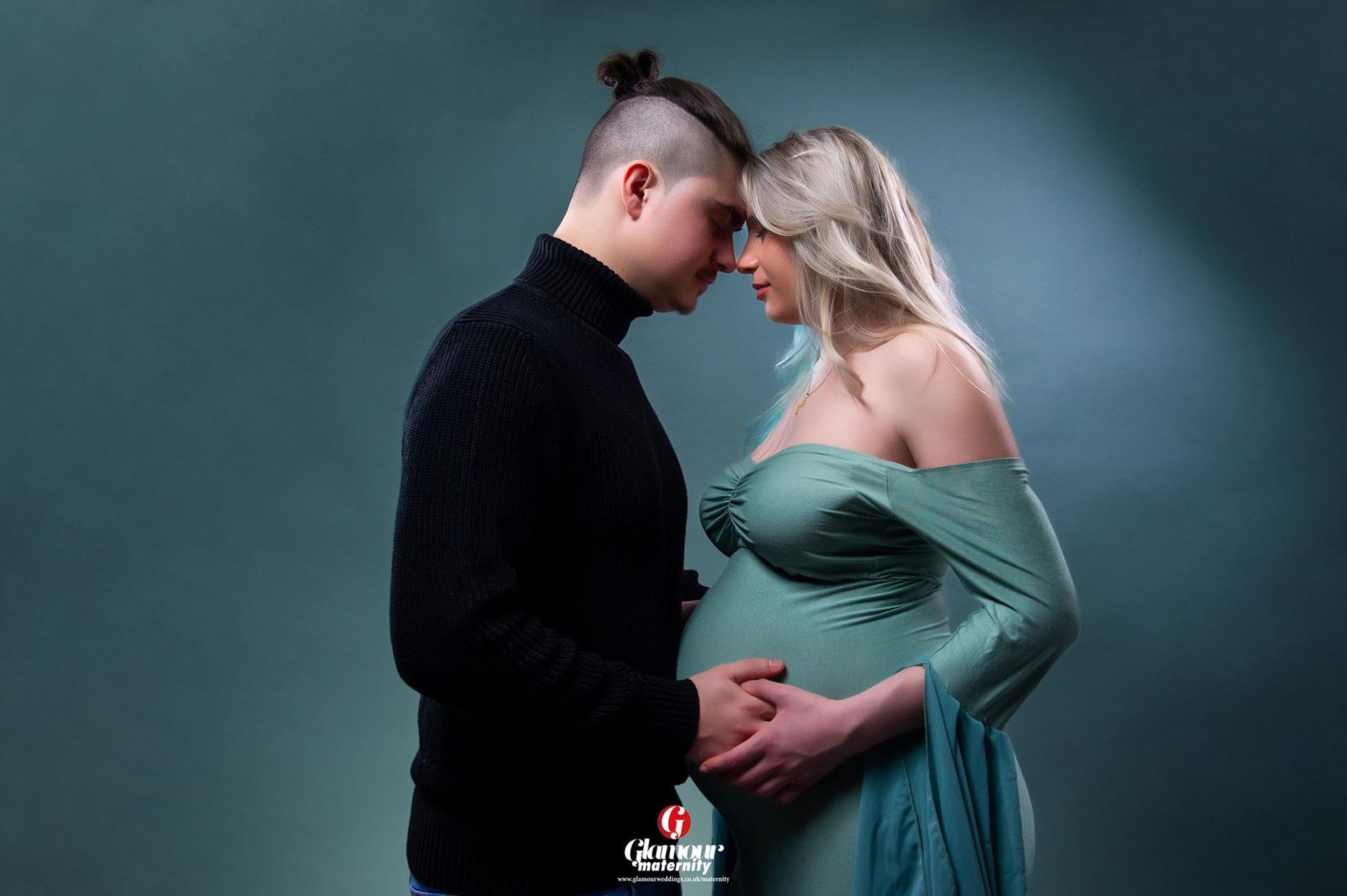 Tender studio maternity portrait of an expectant couple standing closely together in soft sage styling against a teal background.