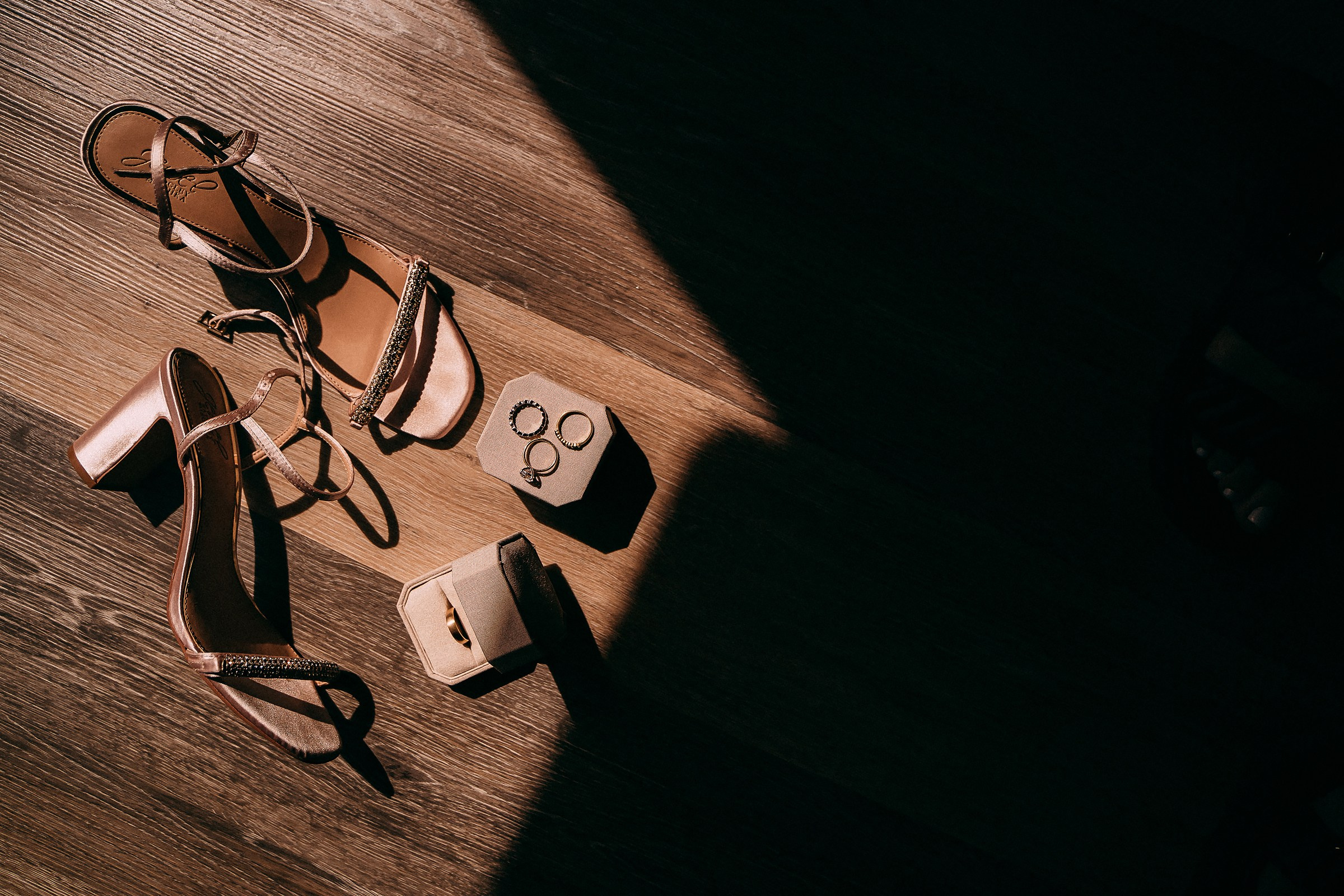 A moody arrangement of wedding accessories including golden rings and strappy heels illuminated by a soft ray of sunlight on a wooden floor.