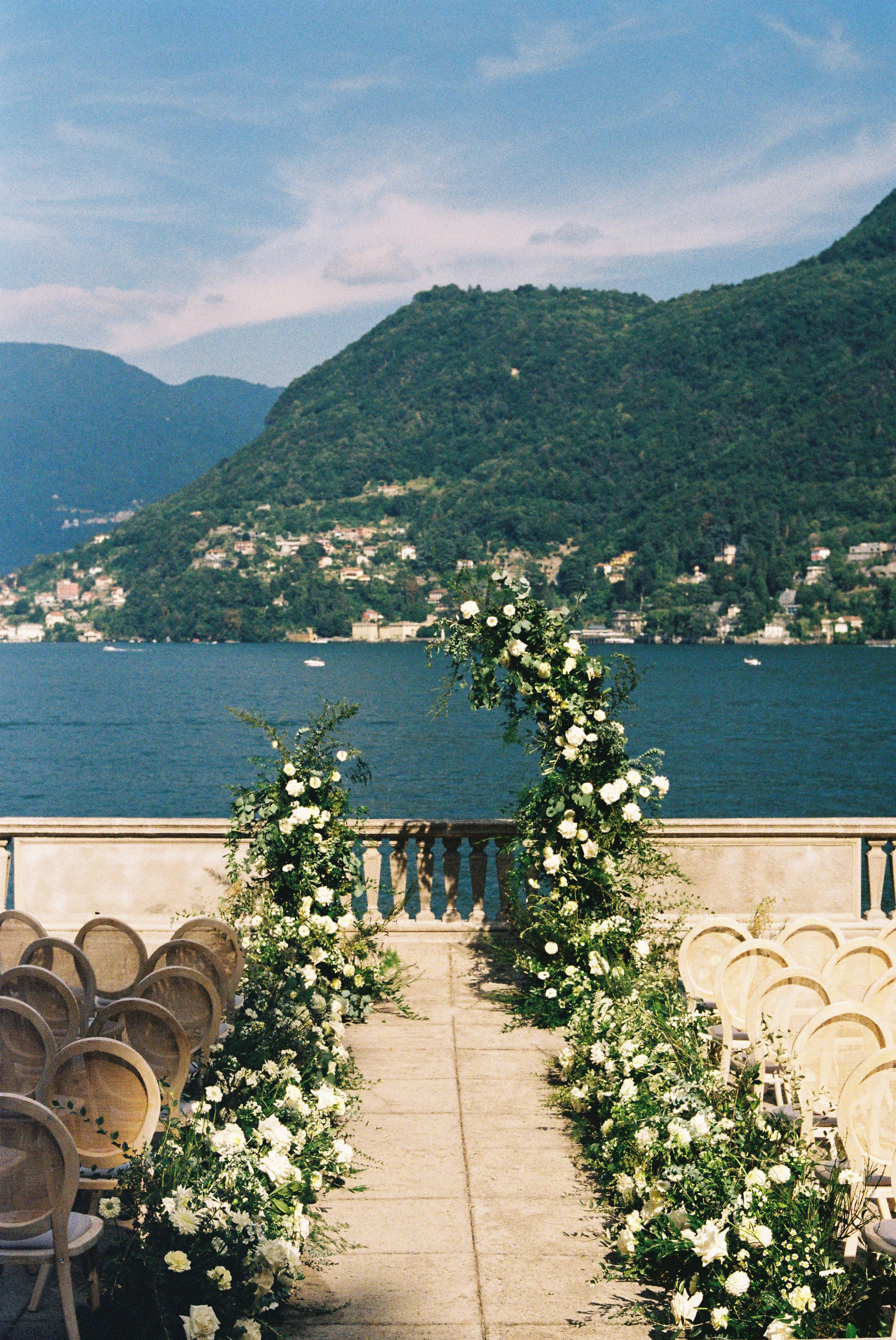 Outdoor wedding aisle with white flower arches, overlooking a scenic lake and mountains.