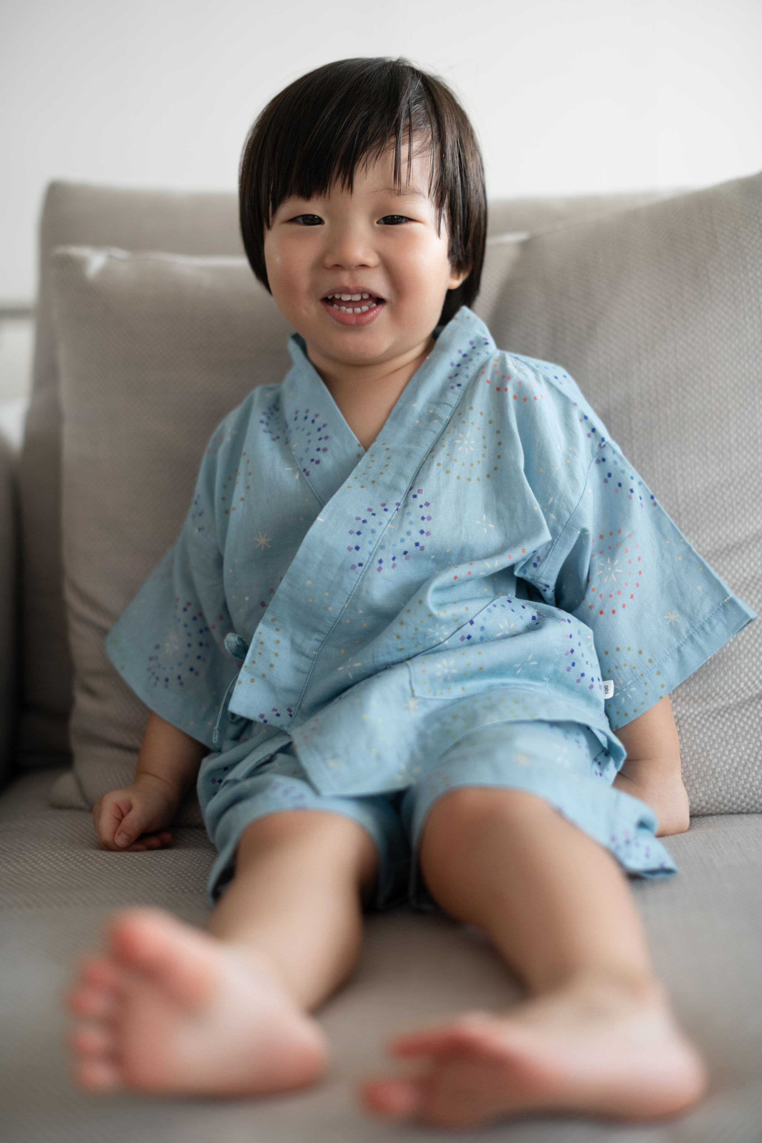 Toddler in a sky-blue yukata smiling and lounging on a beige sofa during a home lifestyle session.