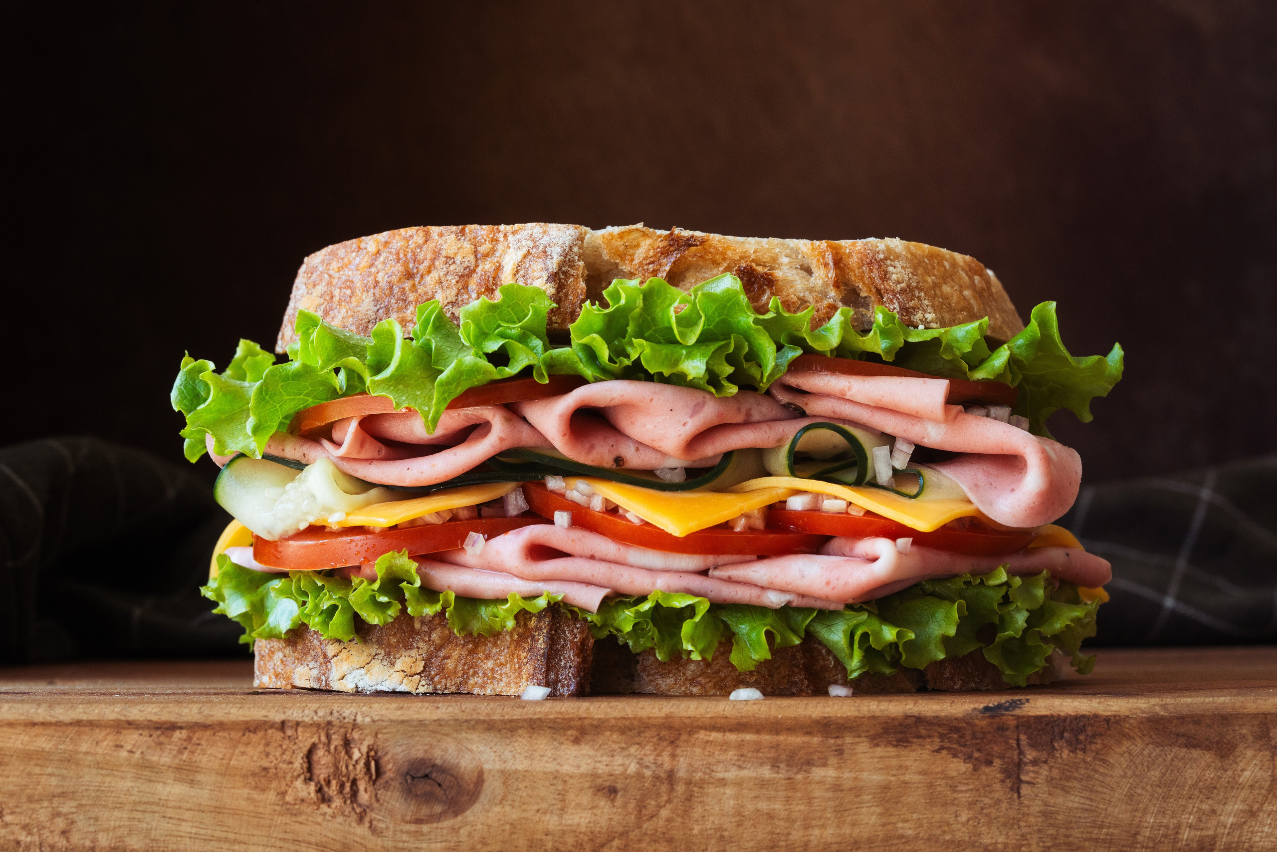 Grande plano de uma grande sanduíche de charcutaria empilhada com fiambre, queijo, alface, tomate e pepino em pão rústico.