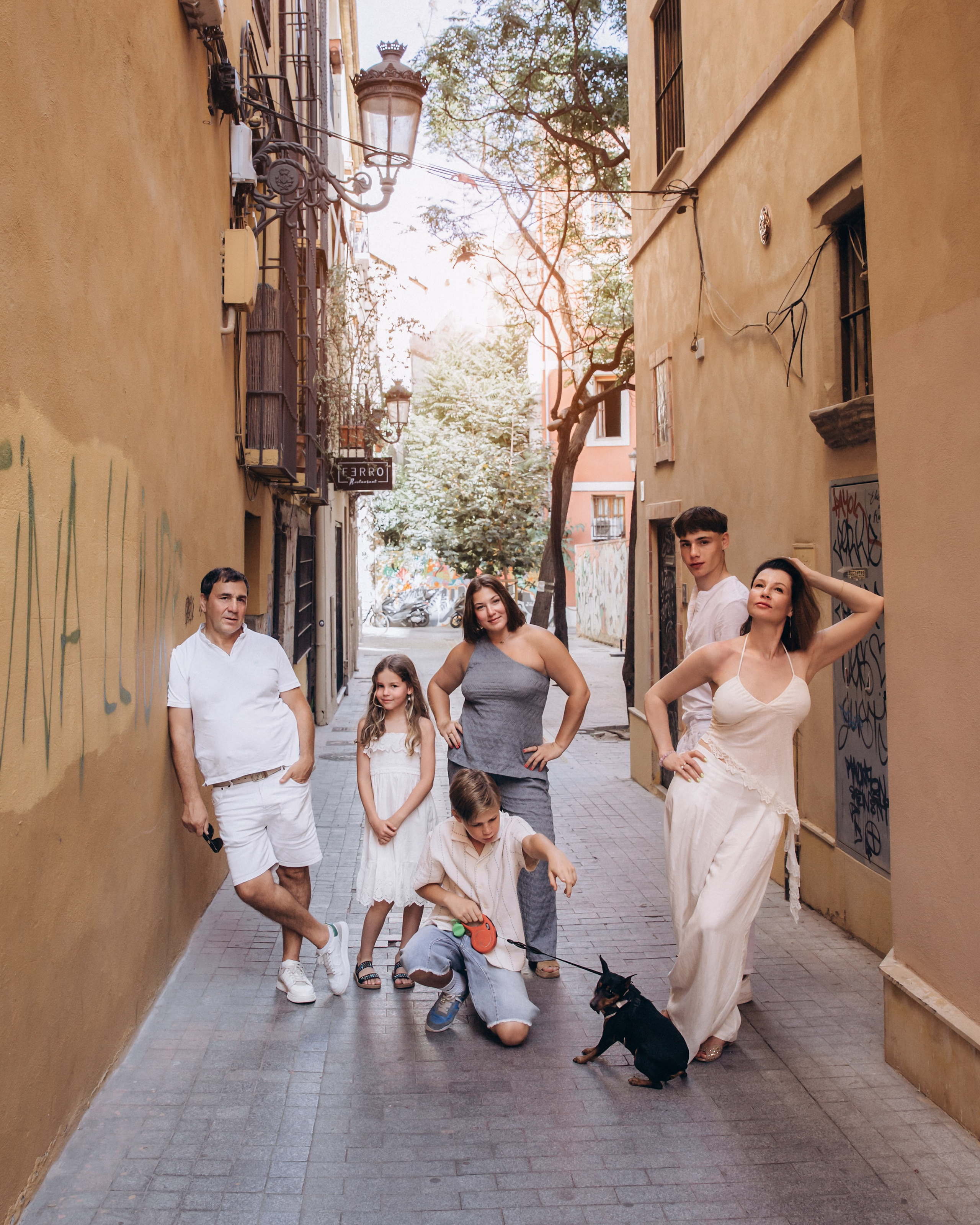 Sesión de fotos familiar en València, España, que captura a una familia elegante y alegre con niños y un perro pequeño posando juntos en una estrecha calle histórica del casco antiguo, rodeados de arquitectura mediterránea cálida y luz natural suave, creando una atmósfera lifestyle y espontánea — ideal para fotografía familiar, sesiones familiares urbanas, retratos familiares lifestyle y sesiones familiares auténticas en València y en toda España.