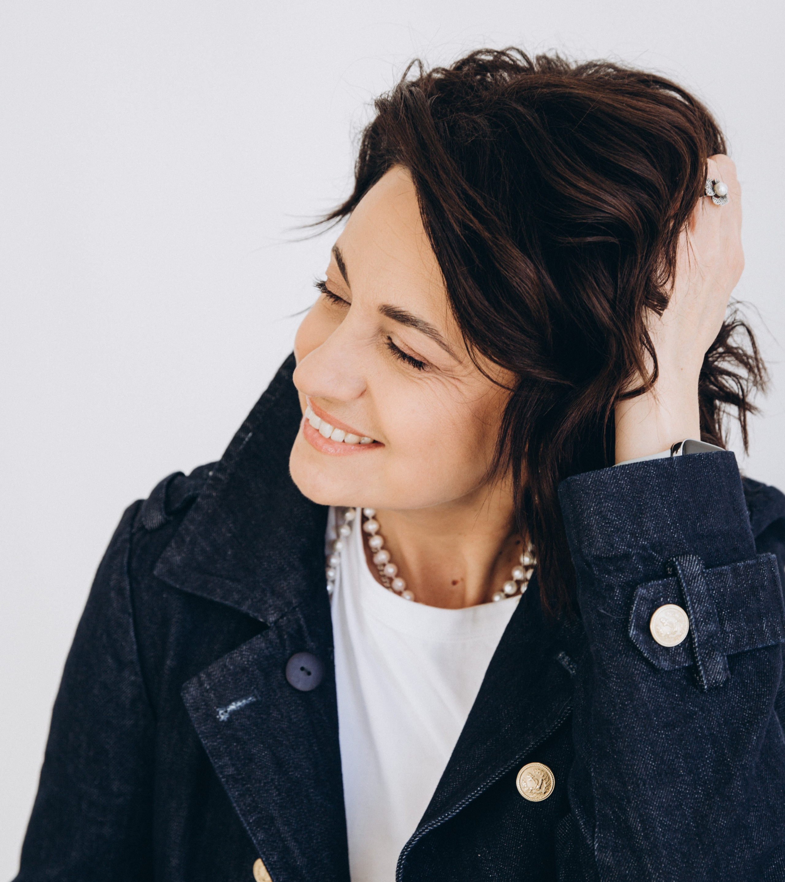 Улыбающаяся женщина в темно-синем джинсовом жакете с жемчужным ожерельем и белой футболке, откидывающая волосы назад в светлой студии в Валенсии, Испания