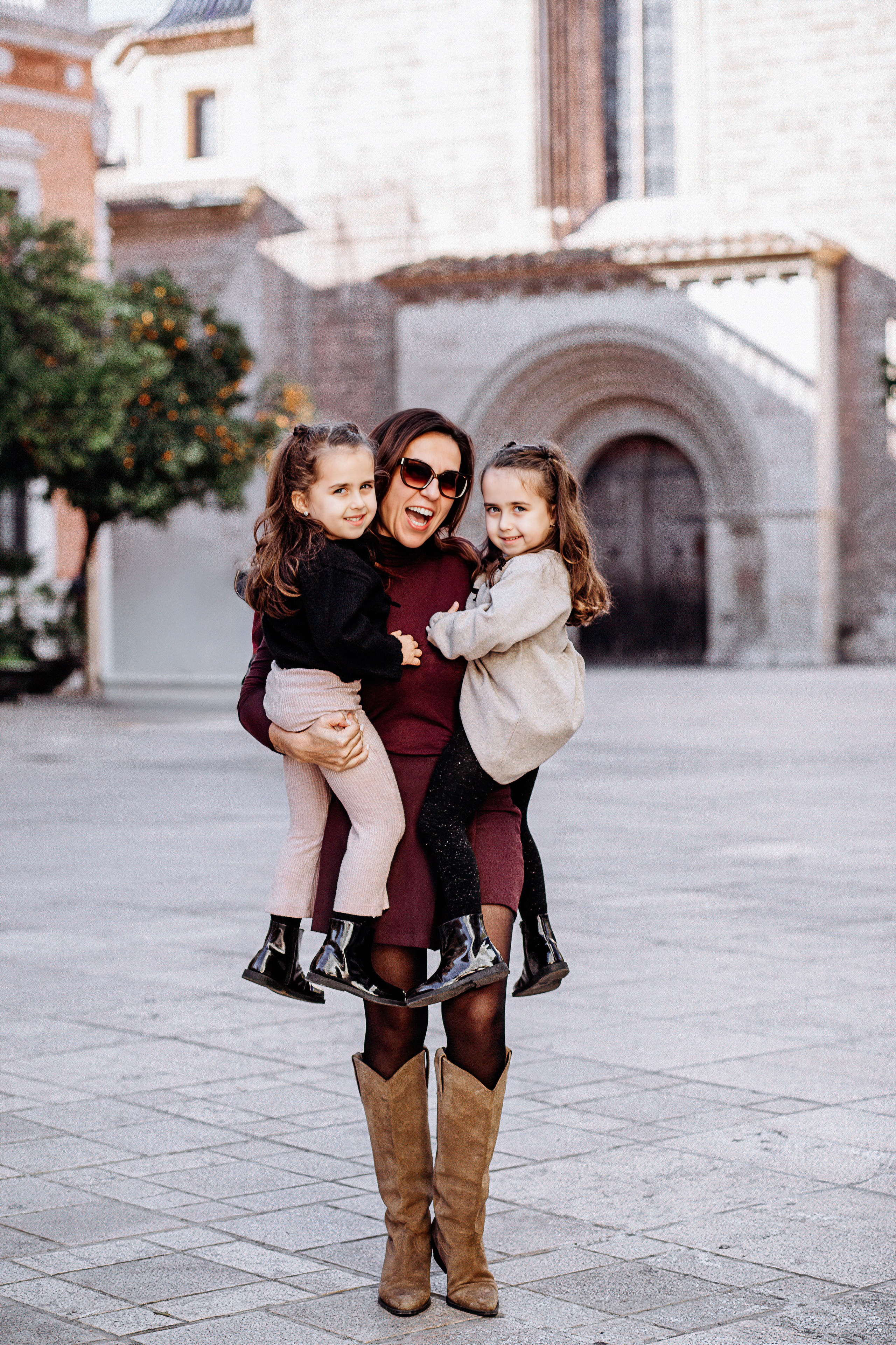 Семейная фотосессия в Валенсии, Испания, где мама держит двух маленьких дочерей в исторической городской локации с очаровательной средиземноморской архитектурой, снято при естественном свете для радостного, живого и эмоционального семейного момента — идеально для семейных фотосессий в Валенсии и по всей Испании.