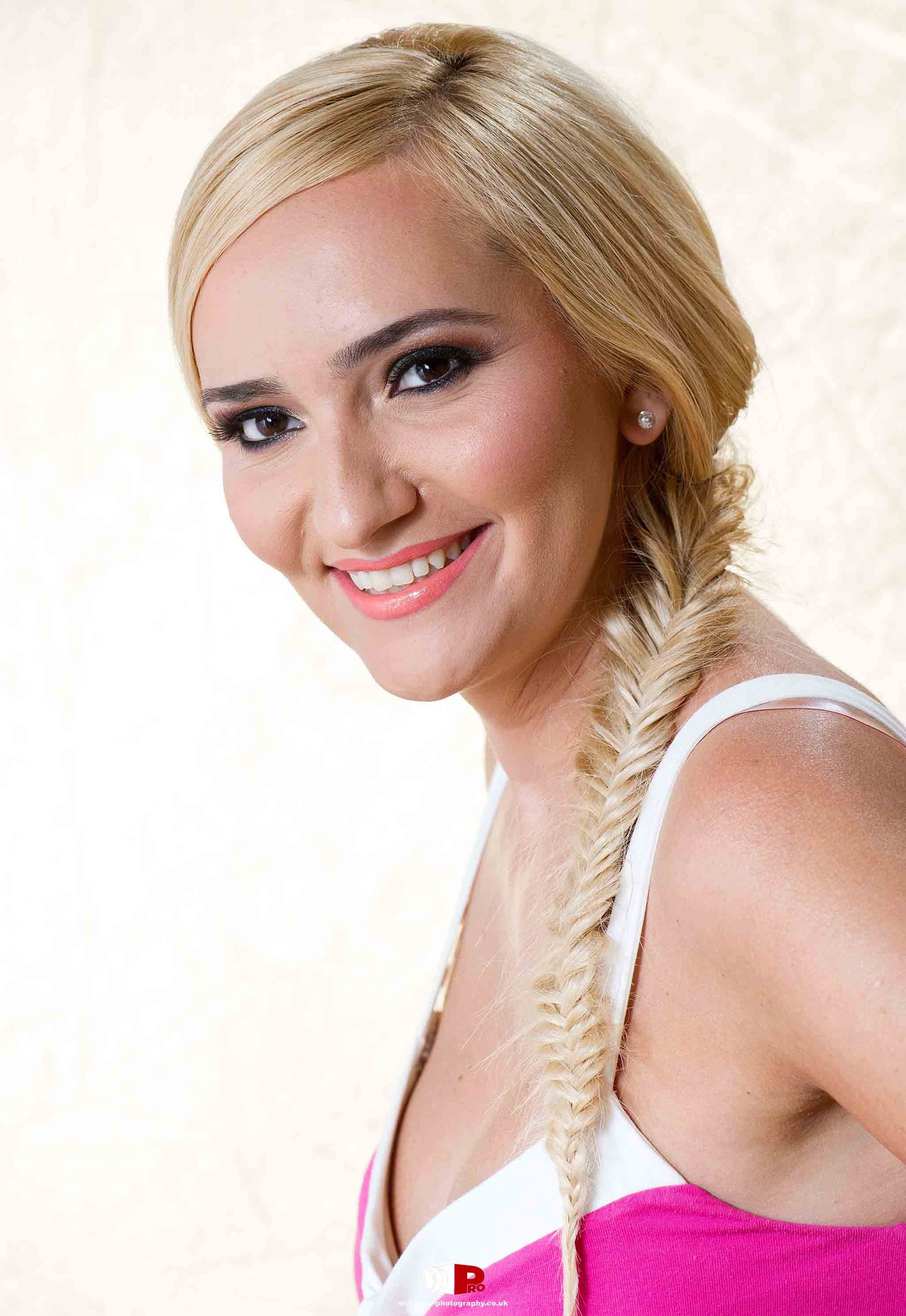 Portrait of a smiling blonde woman with a fishtail braid hairstyle in a pink and white outfit against a soft cream background.