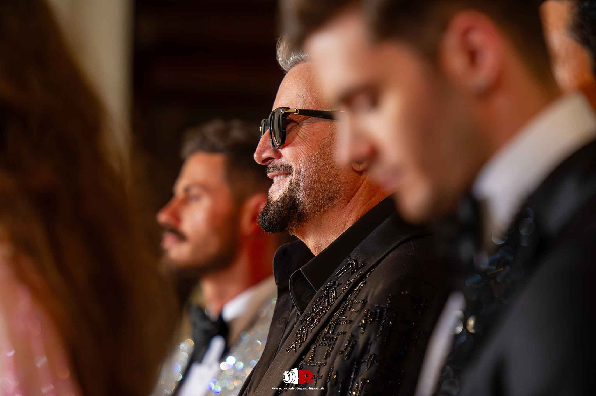 A sharply dressed man with sunglasses smiles confidently while seated among other guests at a fashion event.