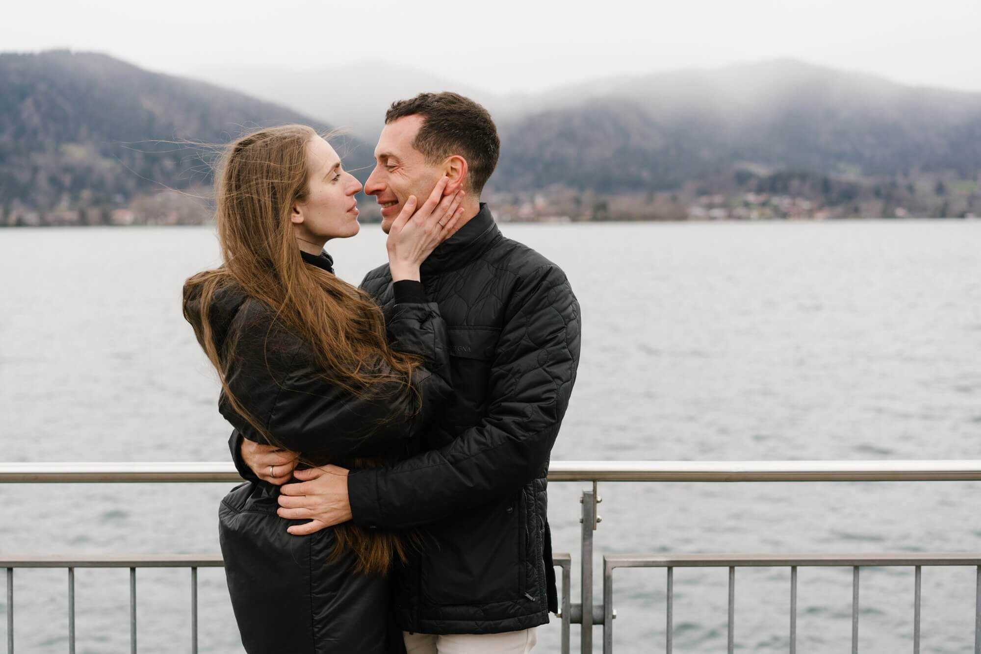 Wide landscape shot of couple at Tegernsee lake dock with misty mountain panorama during couple photo session Germany