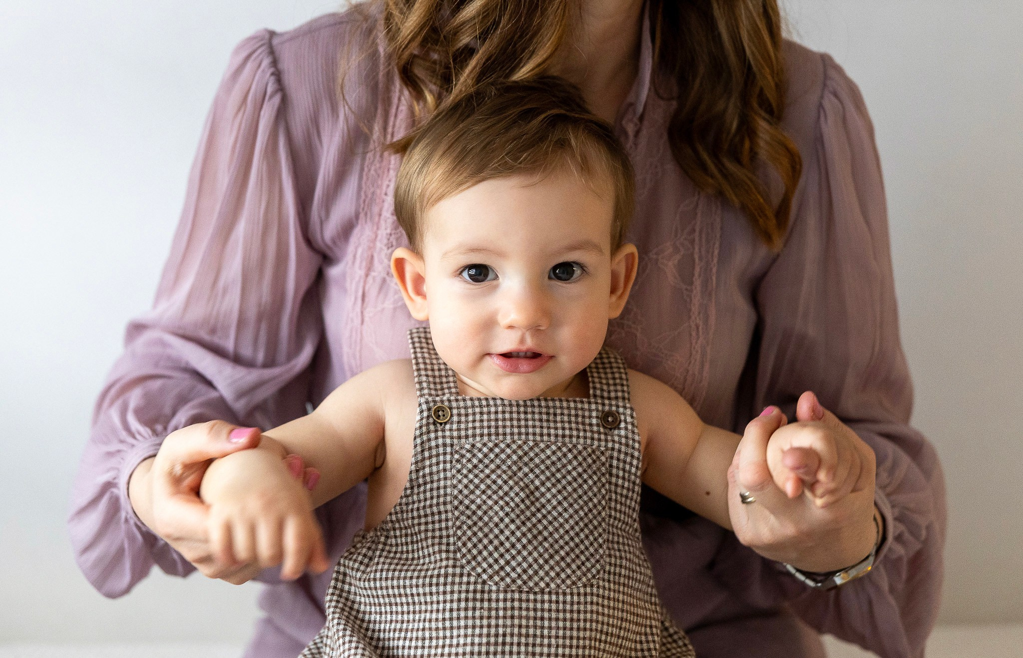 Beba sa smeđom kosom u braon zeki (romperu) sa sitnim kariranim dezenom (gingham), koju drži roditelj u ljubičastoj bluzi. Portret bebe koja gleda u kameru.