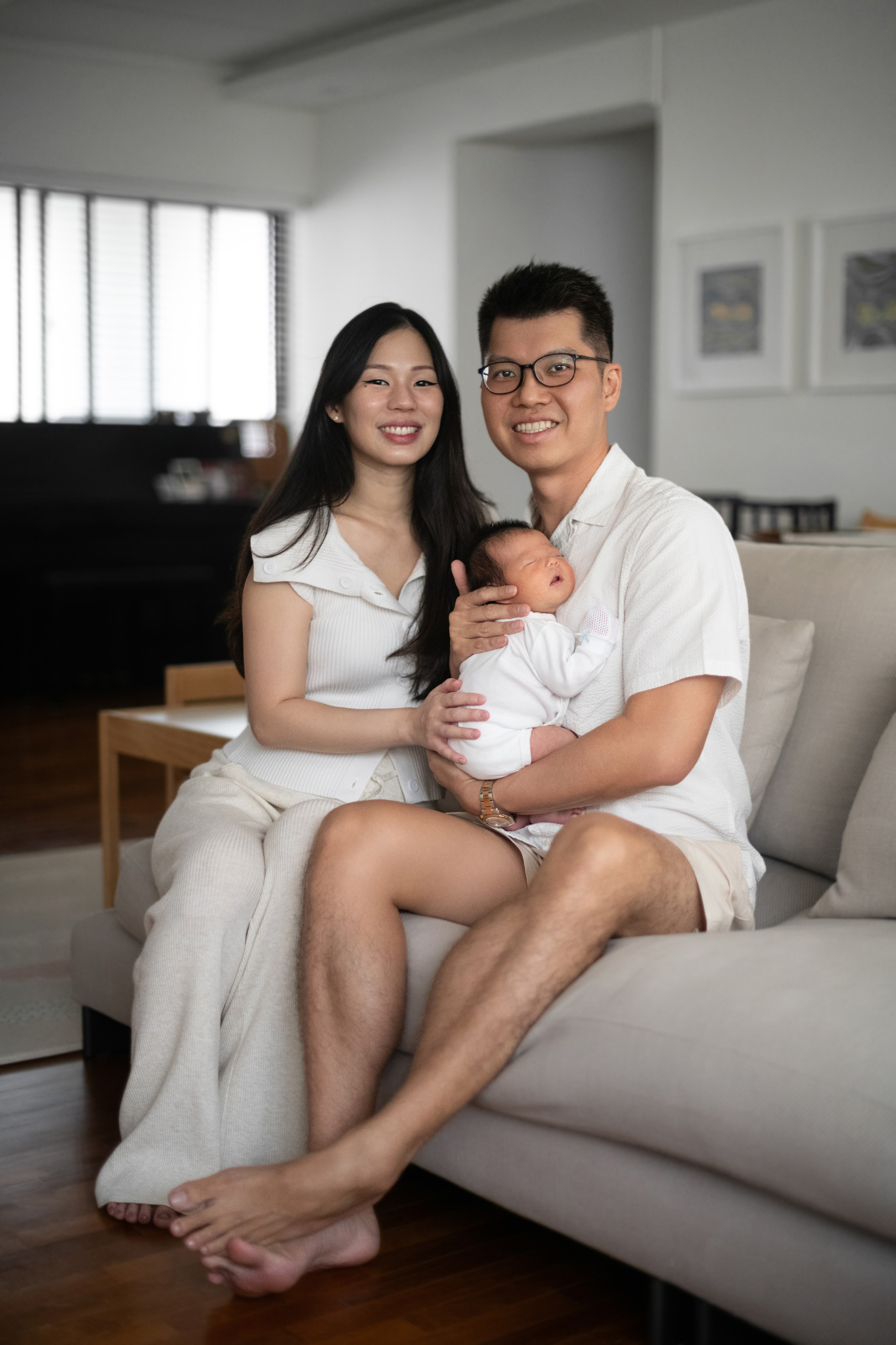 Relaxed family of three on the sofa with newborn, bright living-room scene, candid smiles.