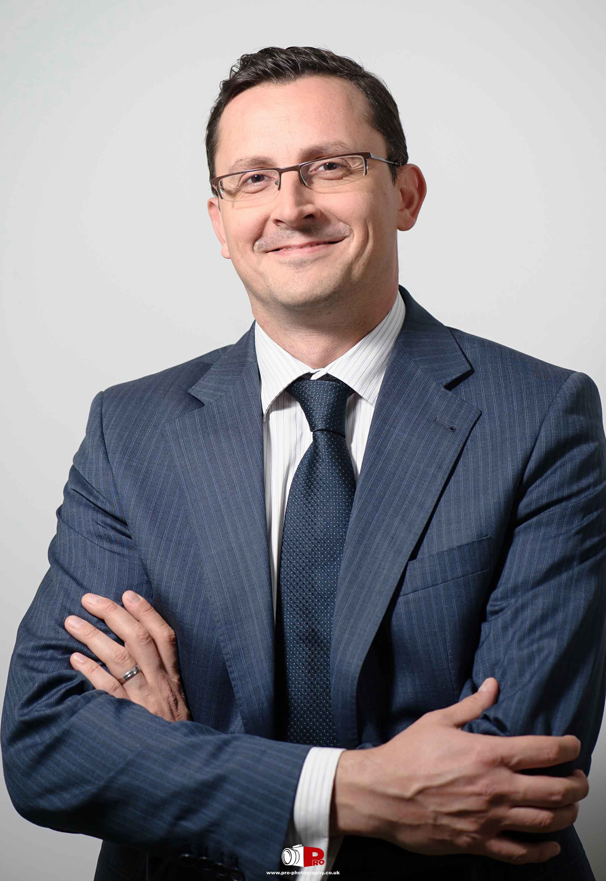 Businessman in a navy blue pinstripe suit with glasses, smiling confidently with arms crossed.