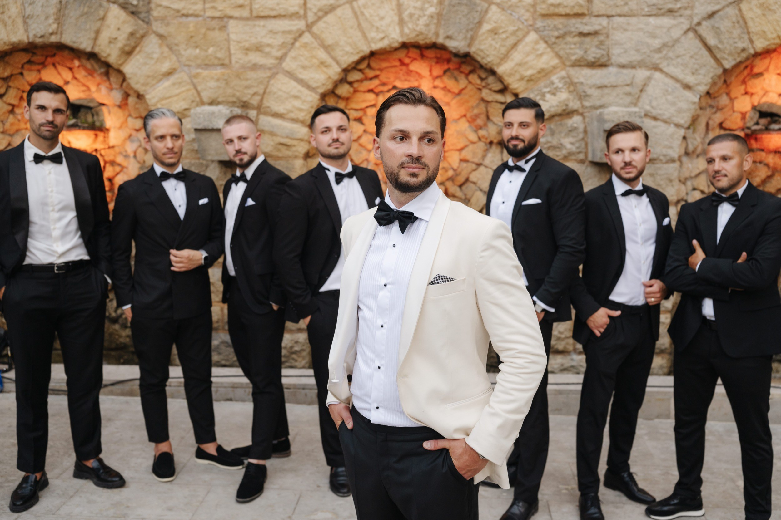 Bräutigam mit seinen Freunden in Smokings am Abend vor einer beleuchteten Steinmauer