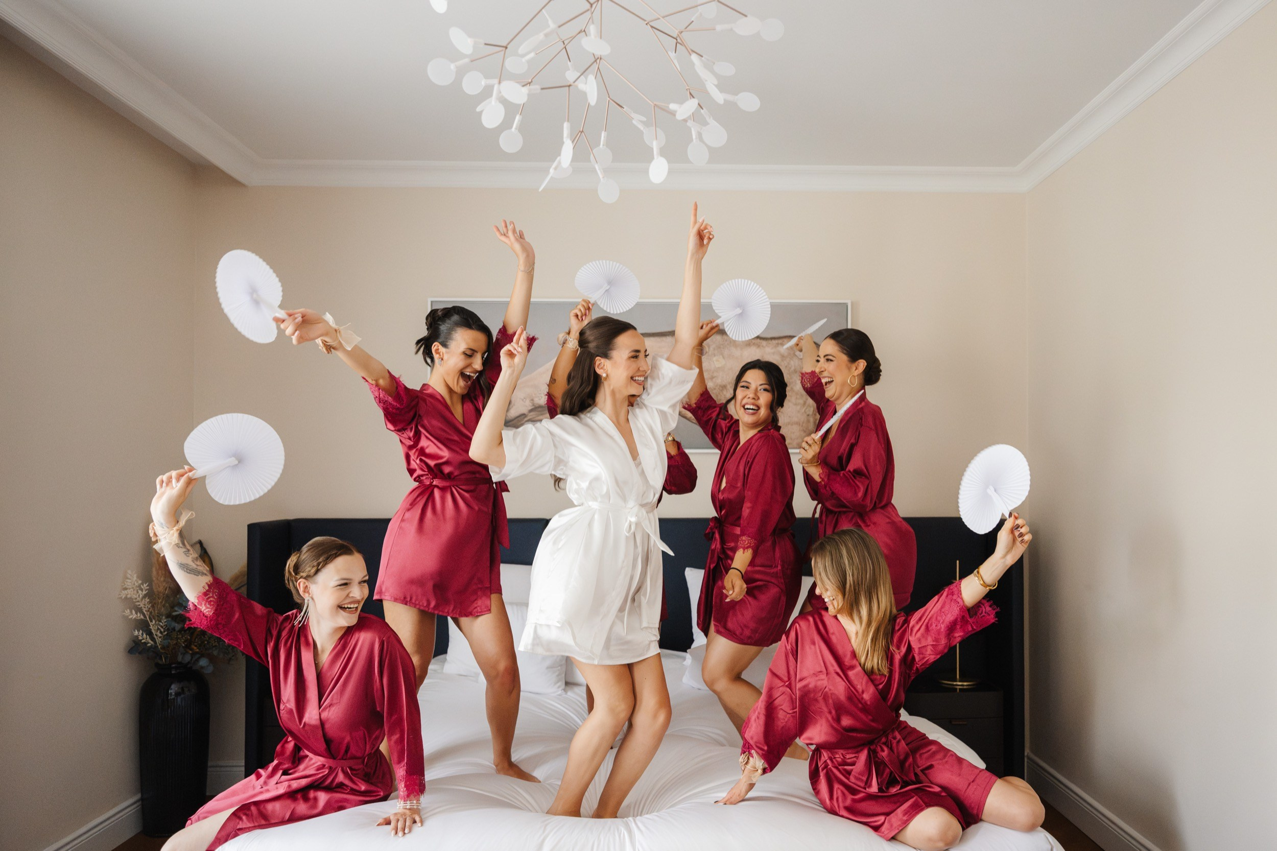 Brautjungfern in roten Morgenmänteln springen mit der Braut jubelnd auf dem Hotelbett