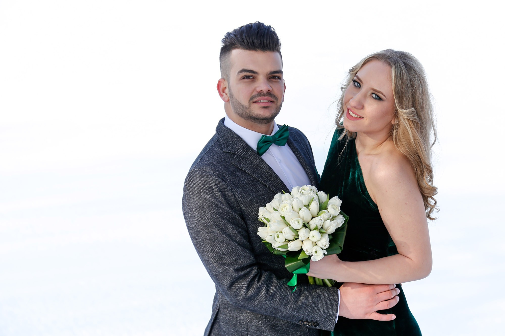 Elegant studio portrait of the newlyweds embracing against a pure white background, showcasing the bride's spectacular emerald green velvet dress.