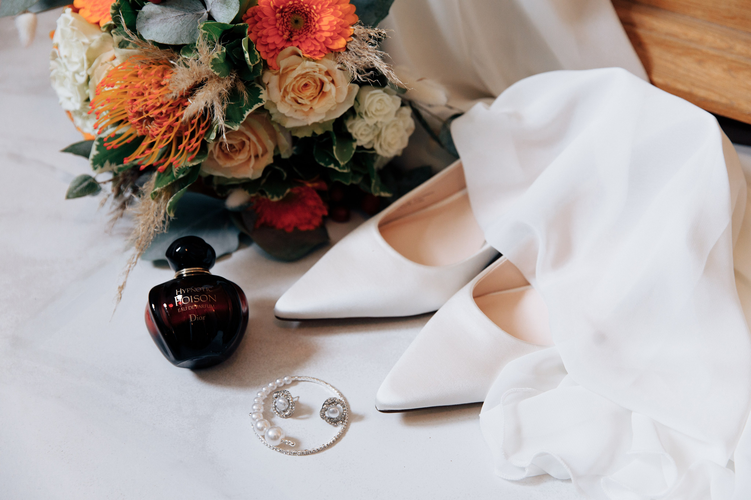 Elegante Hochzeitsschuhe, sanft mit Schleier bedeckt, neben Schmuck, Dior-Parfum und einem Brautstrauß.
