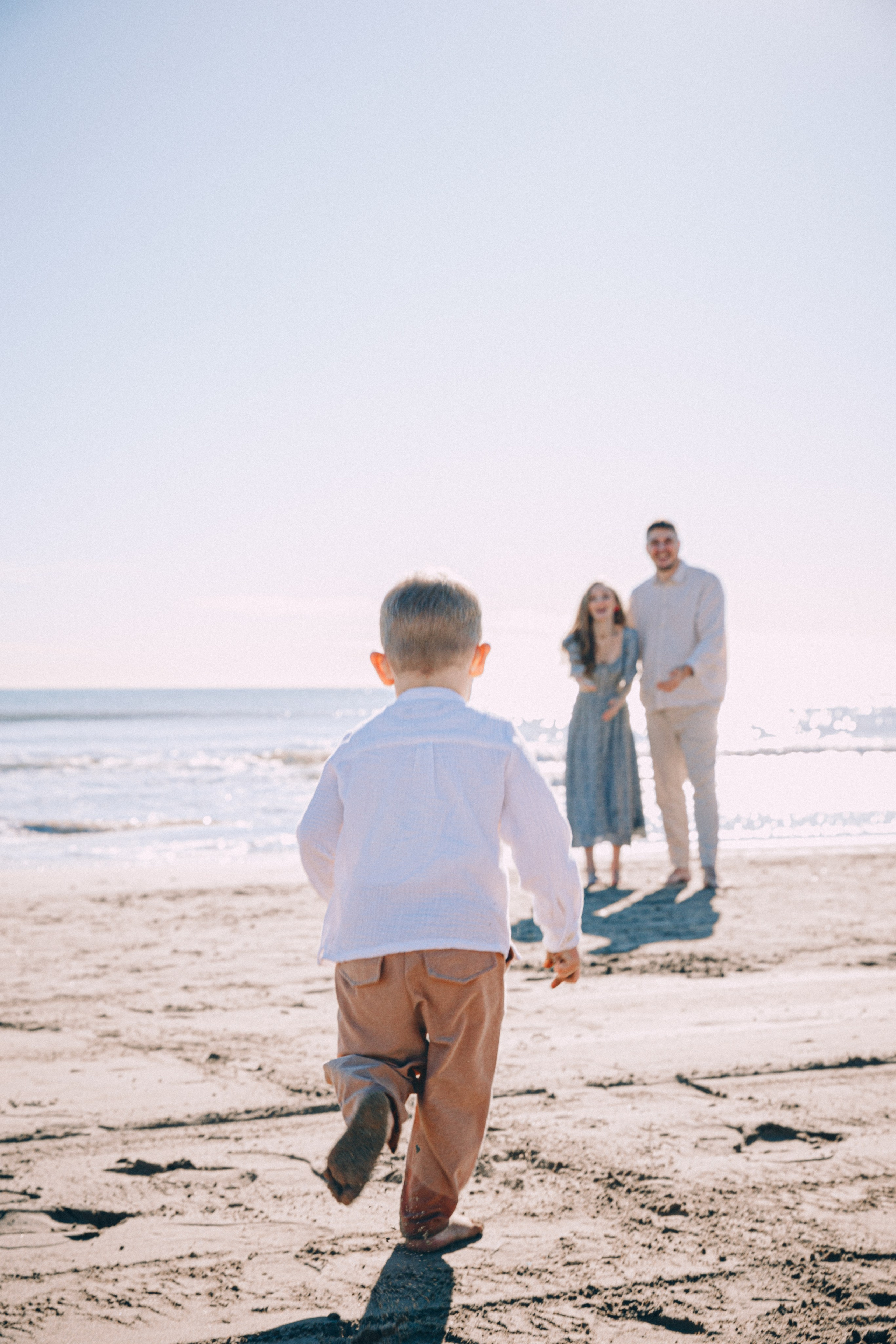 Momento familiar divertido en una playa soleada de Altea, España — un niño pequeño corre alegremente hacia sus padres sonrientes, capturando la calidez y la energía de un día costero sin preocupaciones.