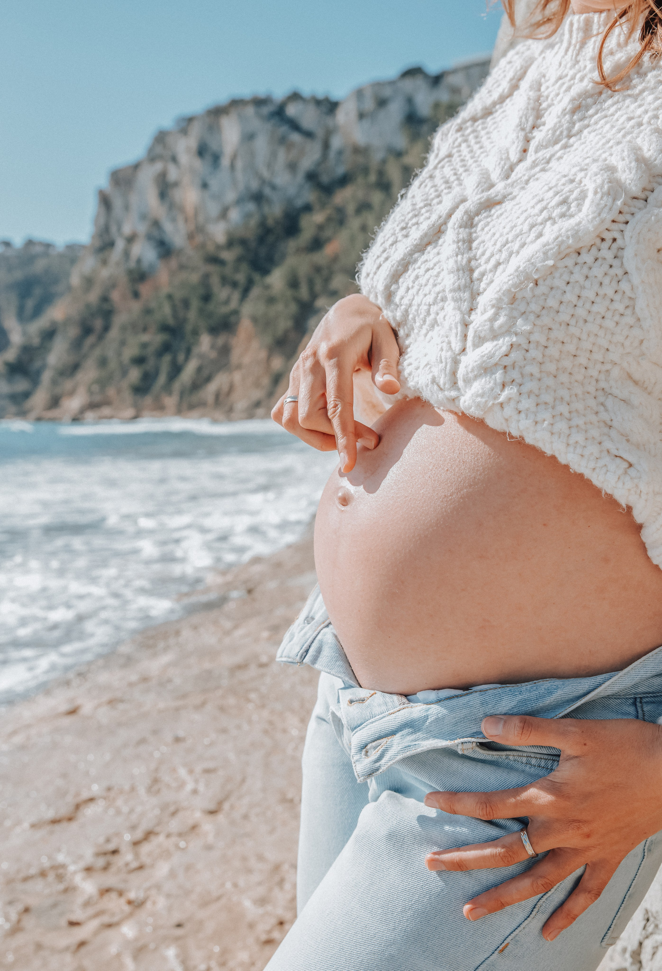 Retrato de maternidad en la playa de Valencia, España — mujer sosteniendo su vientre con las dos manos, vestida con suéter blanco y vaqueros, con acantilados y olas bajo un cielo azul.
