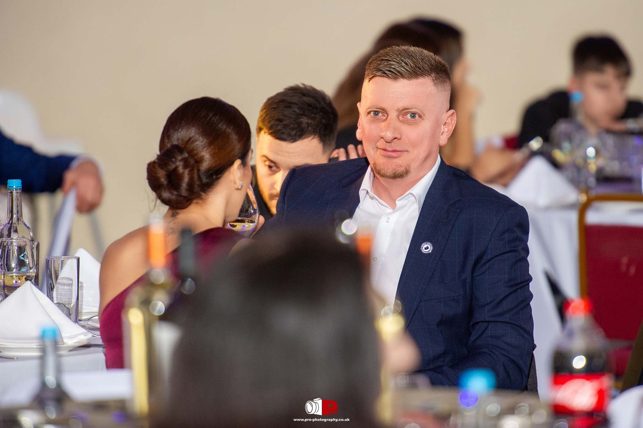 A man in a navy suit enjoys a fine dining experience at a formal evening event with wine and guests in the background.