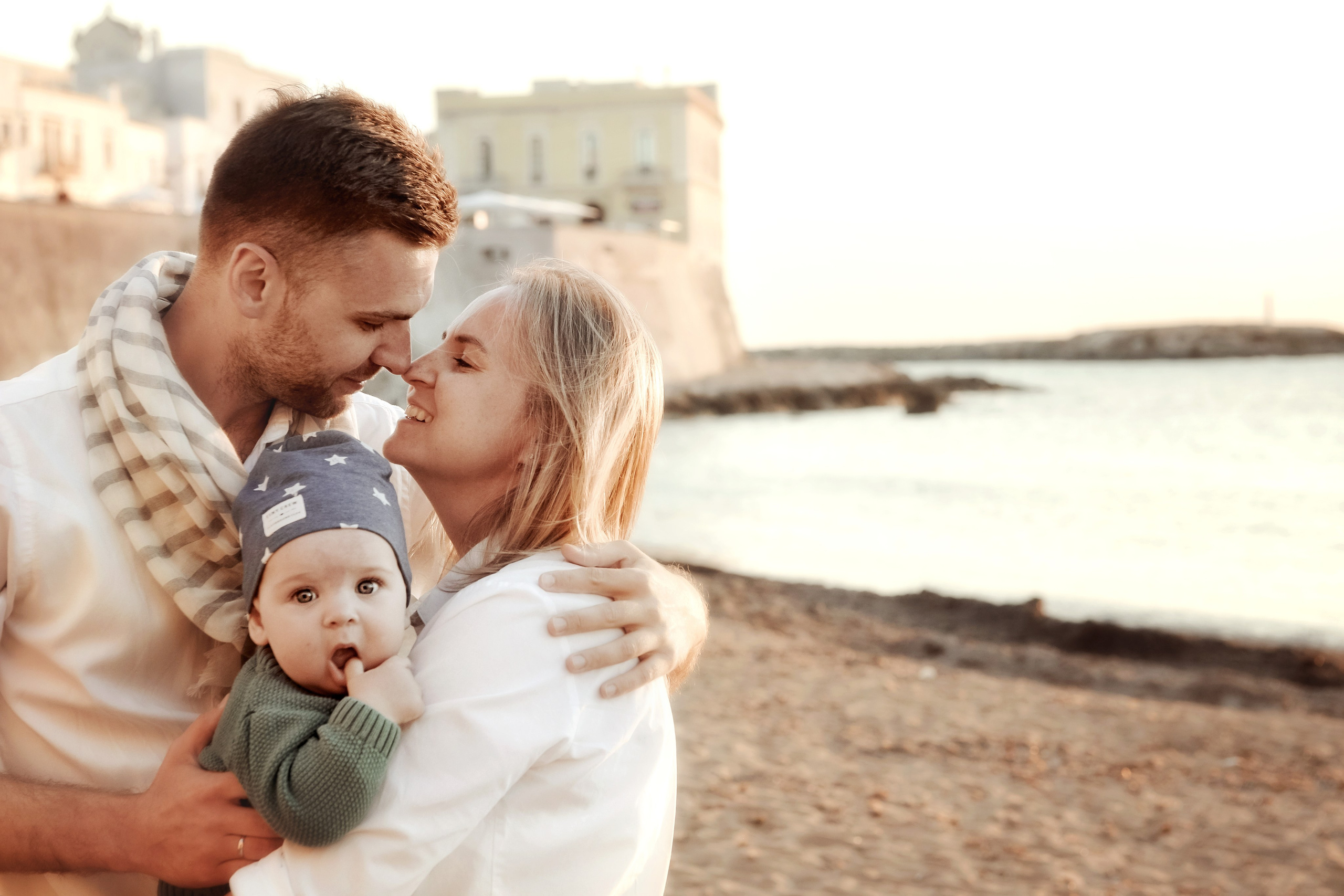 A tender family moment on the beach in Puglia: a young couple embraces with their baby at sunset, bathed in warm golden light.