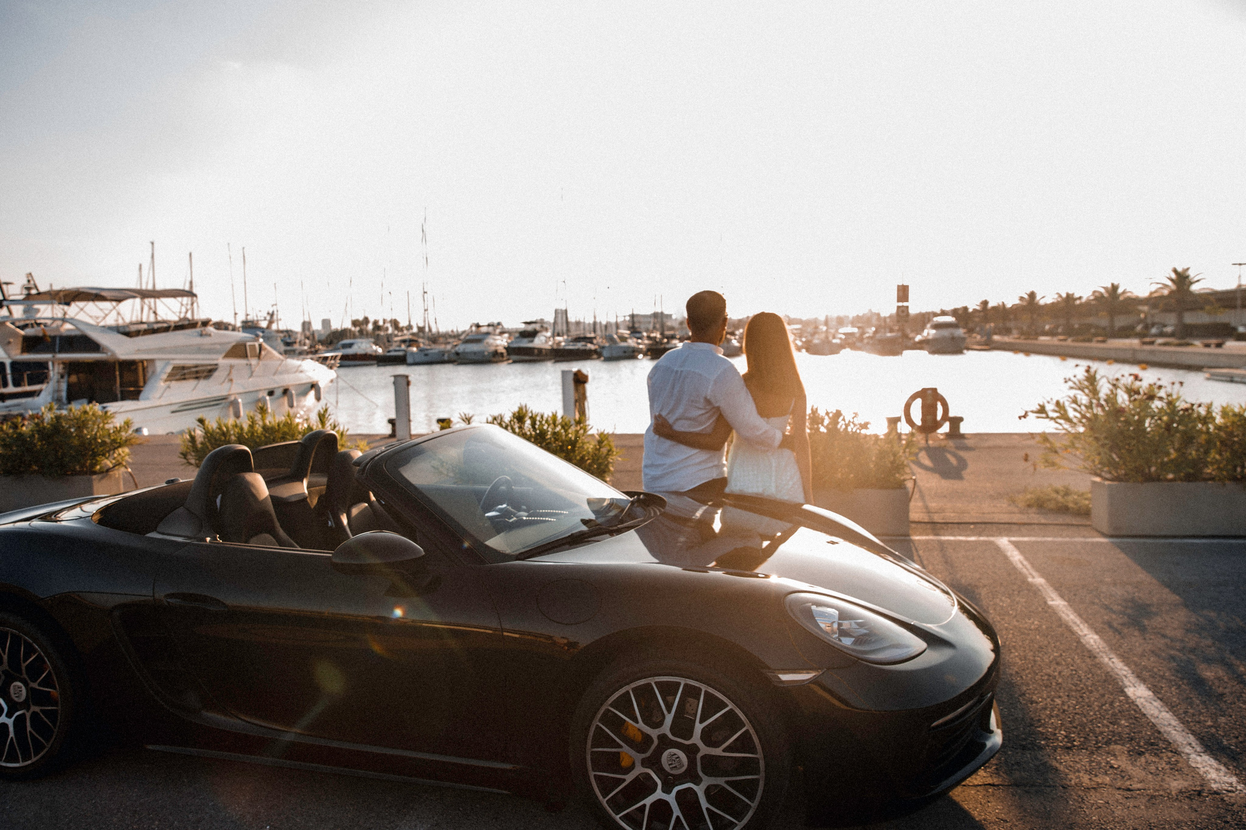 Romantic couple embracing at sunset by the marina, standing near a black convertible sports car with yachts and palm trees in the background in Valencia, Spain — a stylish love story photoshoot capturing luxury and intimacy.