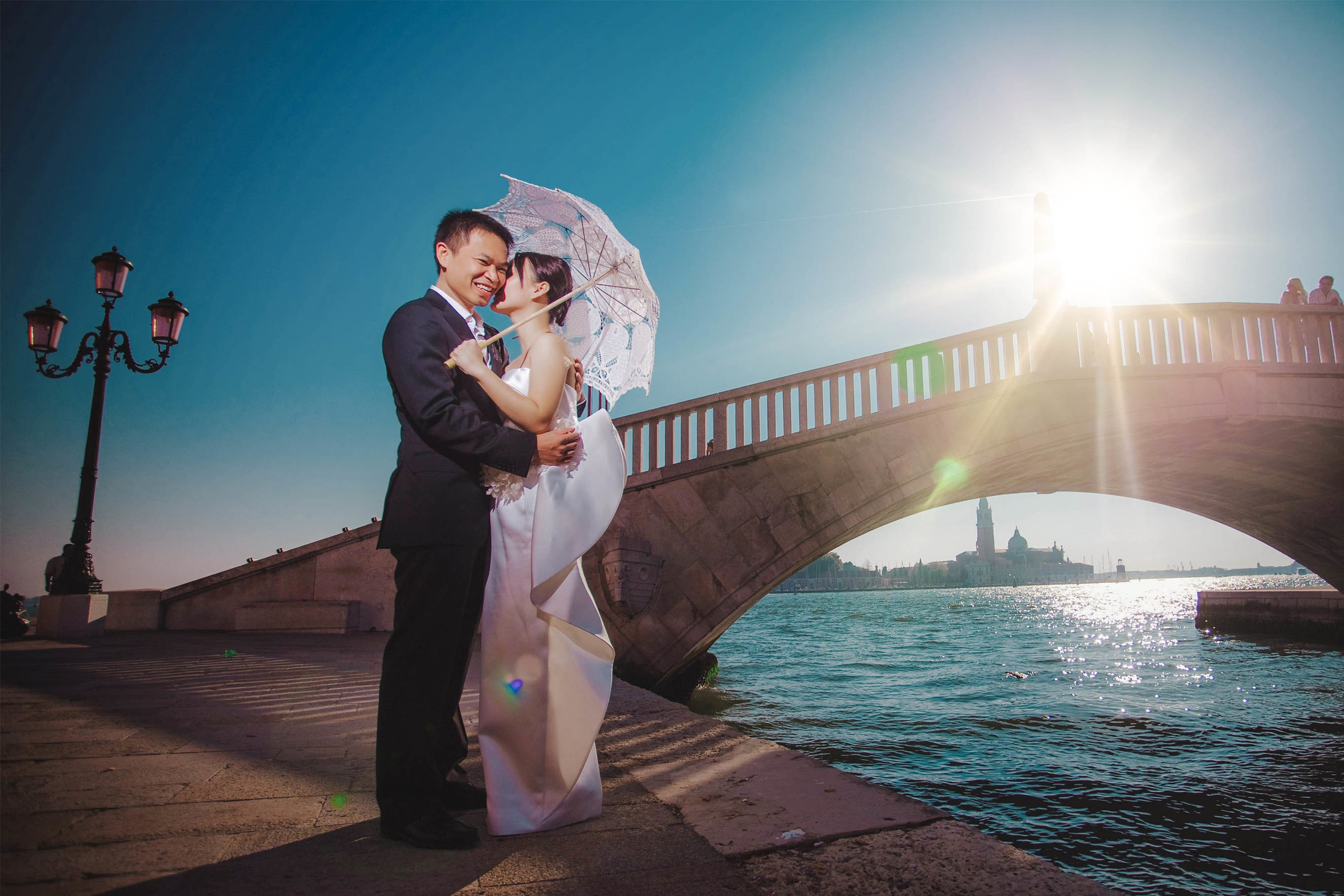 Young smiling Thai bride holding parasol embracing partner near gondolas in Venice.