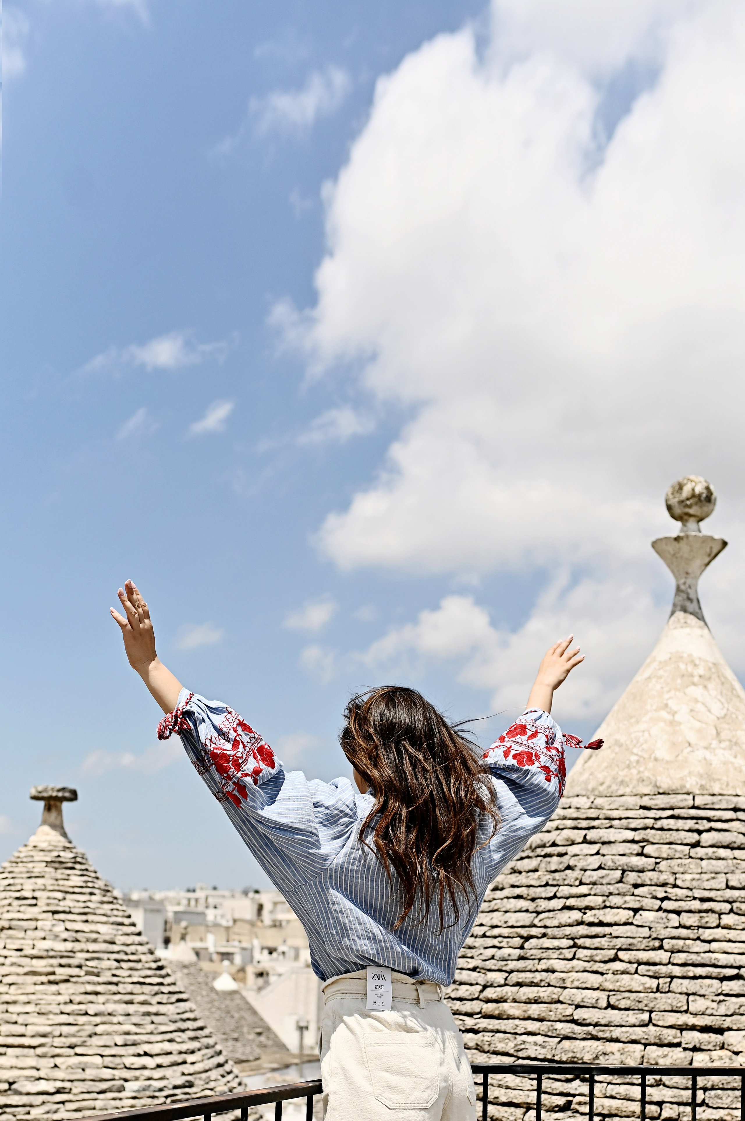 A woman in an embroidered blouse and light shorts raises her arms to the sky, standing on a terrace overlooking the trulli rooftops in Alberobello.