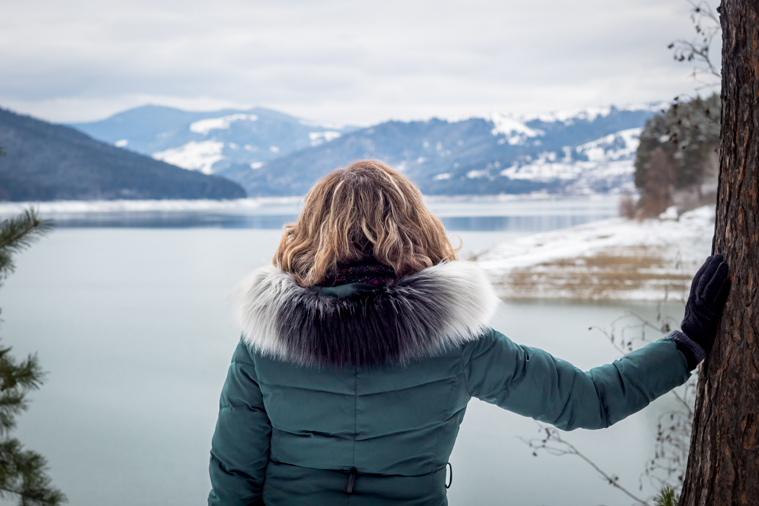 Woman in winter jacket bracing wind with snowy mountains and lake in the background.