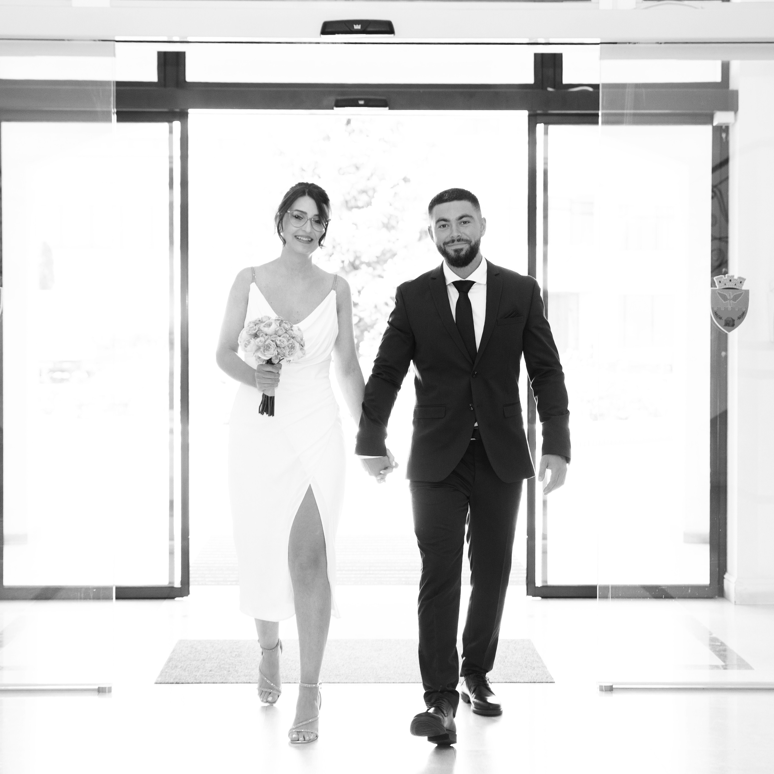 Bride in white dress holding bouquet and groom in dark suit walking through glass doors.
