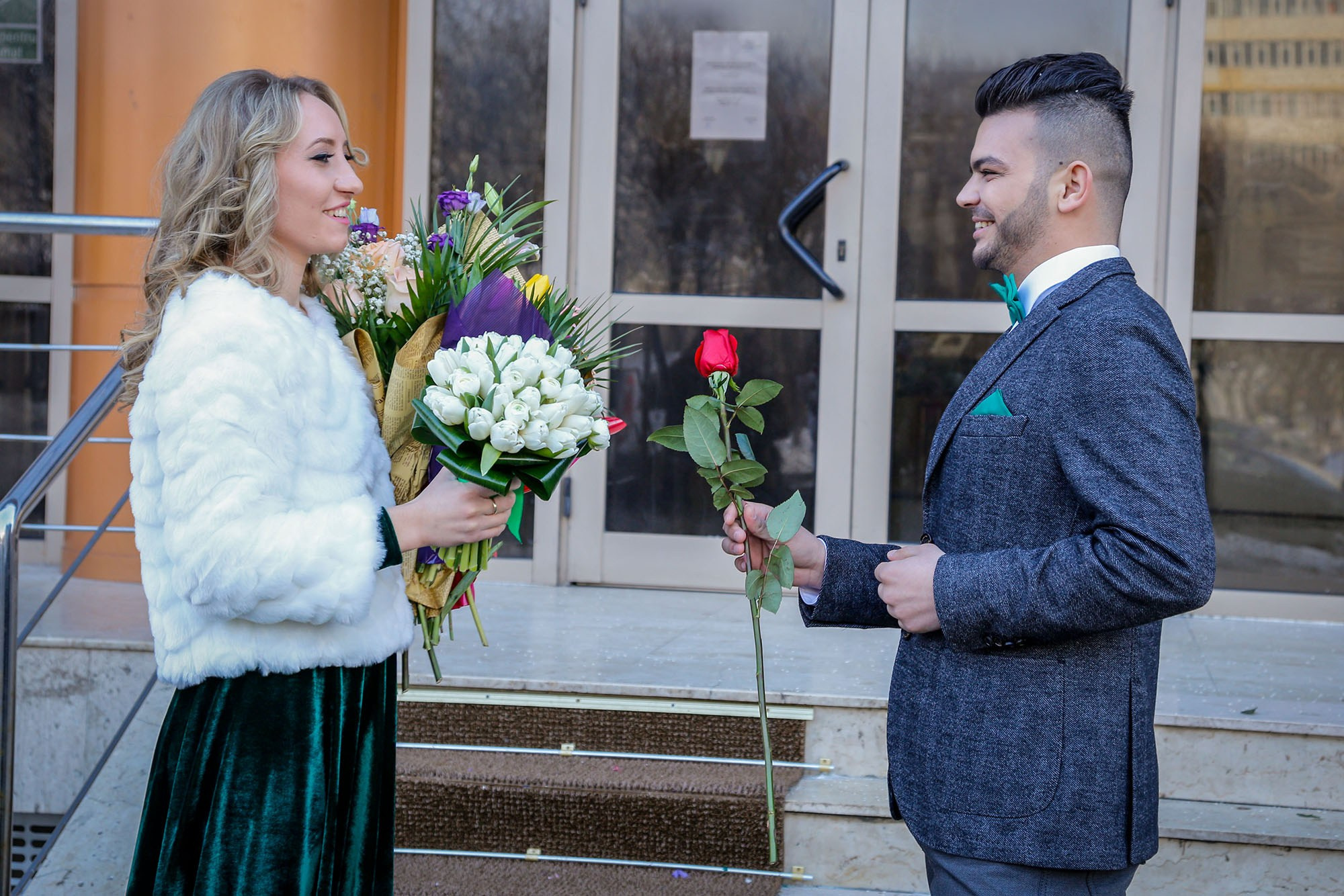 Newlyweds standing face to face outdoors, with the groom offering a beautiful red rose to his bride during their wedding photo shoot.