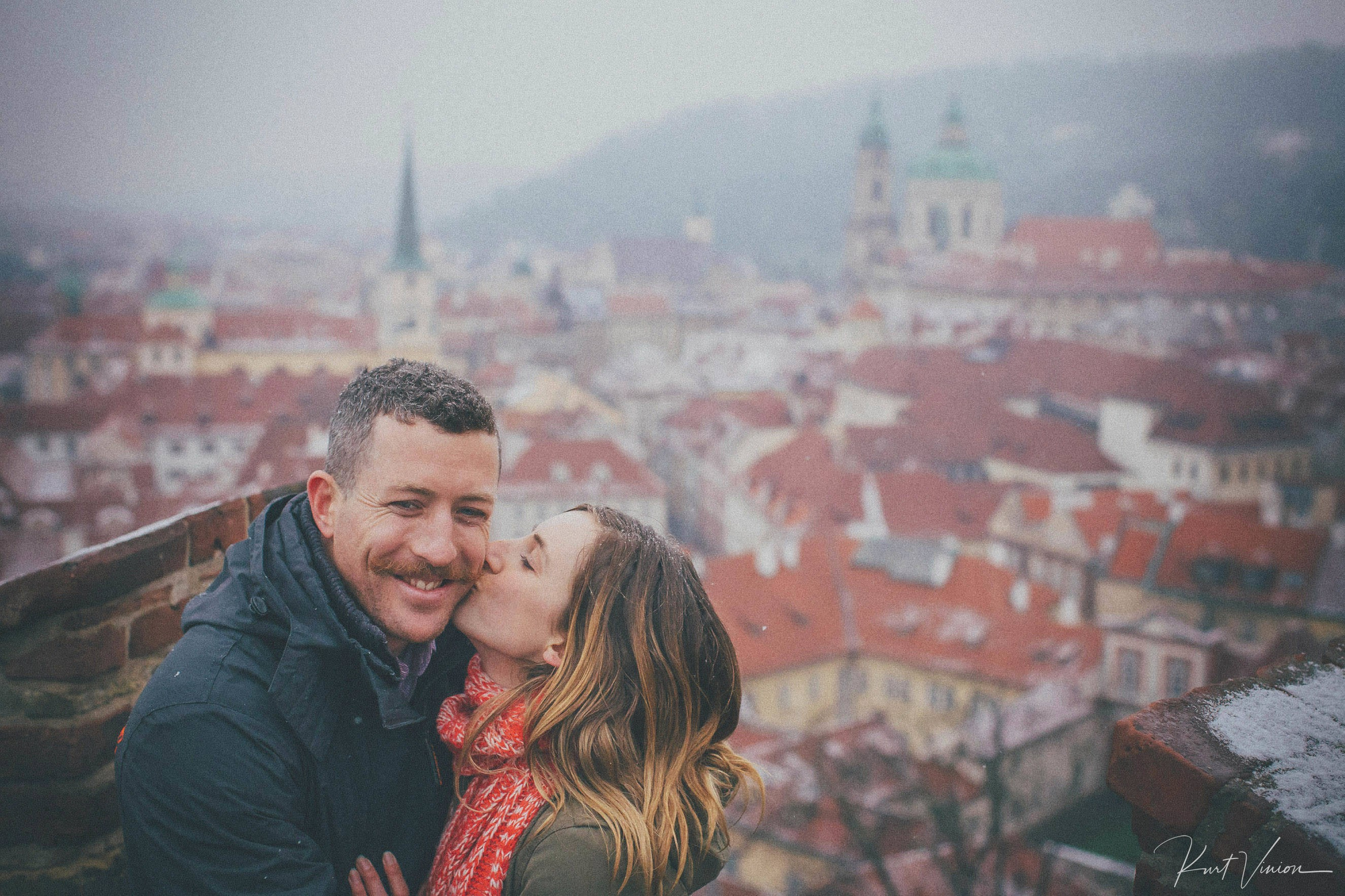 Woman kissing partner on cheek overlooking red-roofed Mala Strana from Prague Castle on winter day.