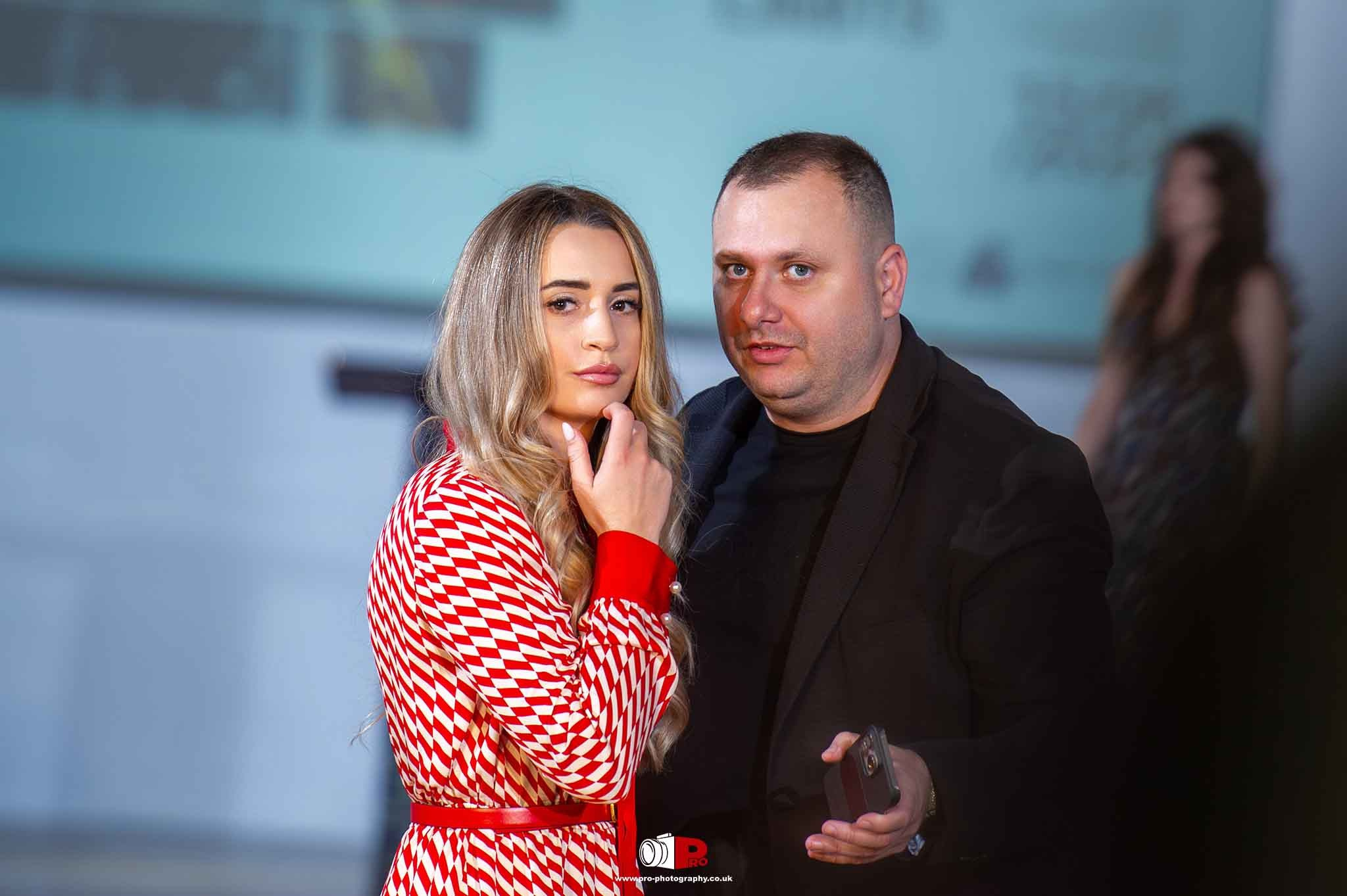 A man and a blonde woman in a red patterned dress discussing event details at a high-profile gathering.