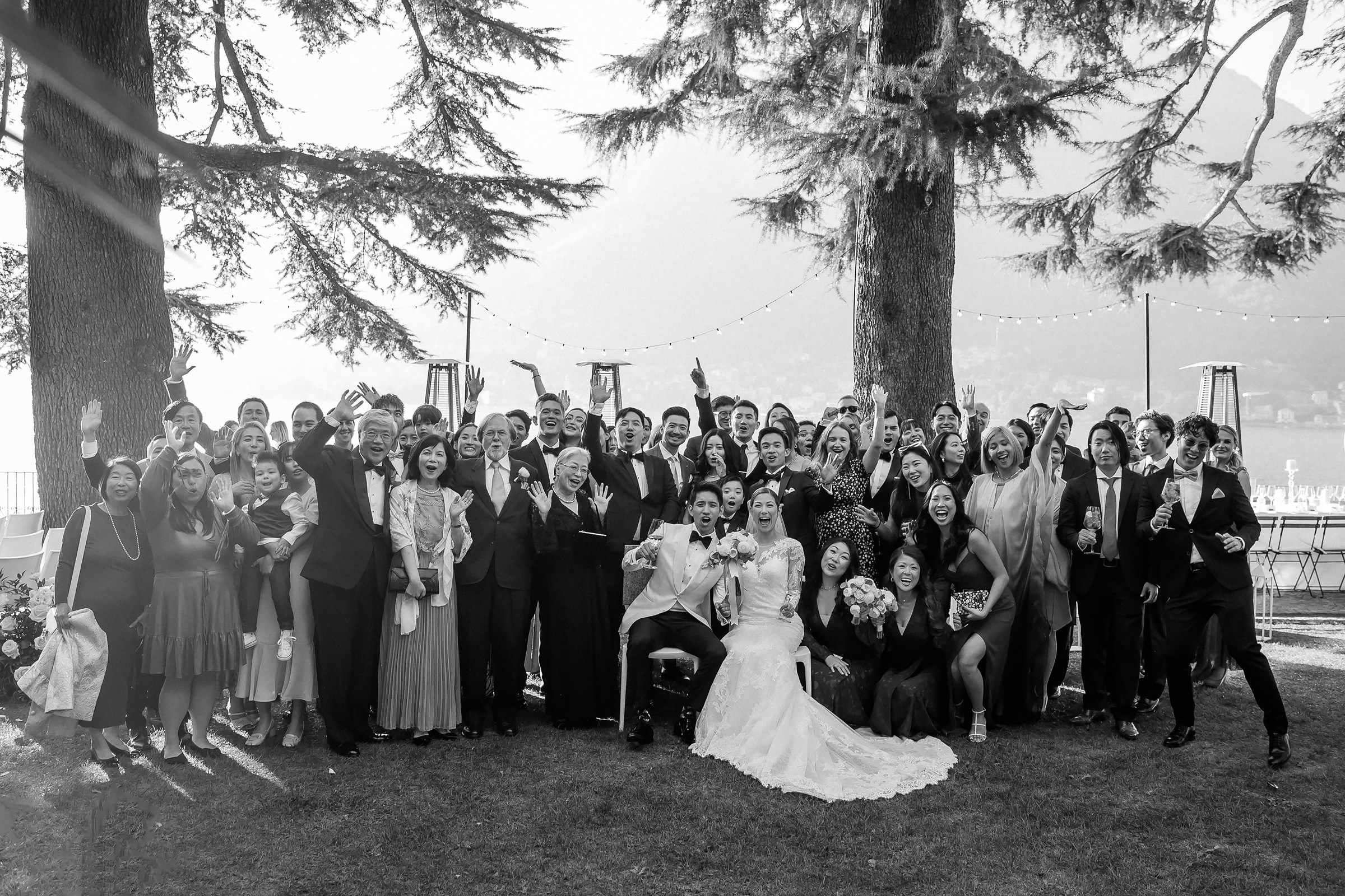 The entire wedding party gathers under large trees, with everyone smiling and raising hands in celebration.