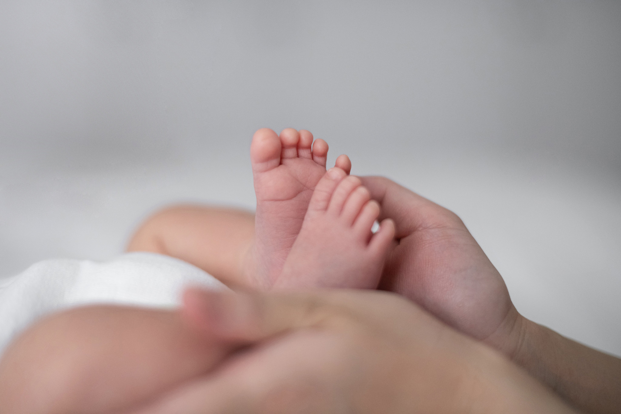 Macro of tiny newborn feet cupped in mum’s hands against a soft grey backdrop, shallow depth of field. 	