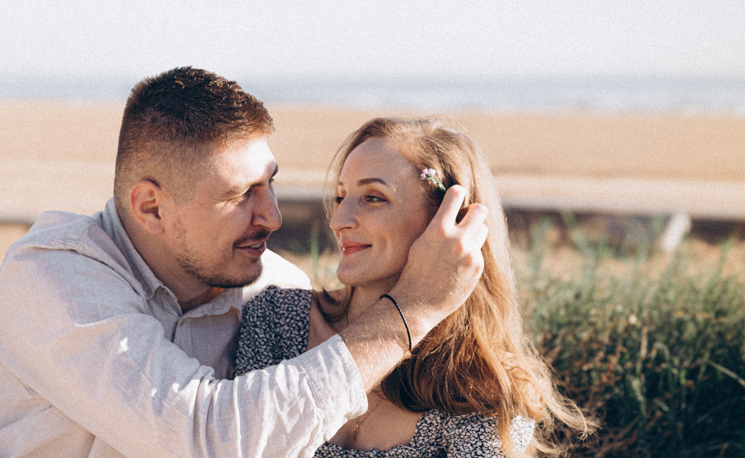 Sesión de fotos love story de pareja en València, España, capturando un momento íntimo y emocional en el que la pareja comparte una mirada tierna, mientras el hombre acaricia suavemente el cabello de su pareja, fotografiado en exterior con luz natural suave y un cálido fondo mediterráneo — ideal para fotografía love story, sesiones de pareja, reportajes de compromiso, retratos lifestyle románticos y fotografía de pareja en València y en toda España.