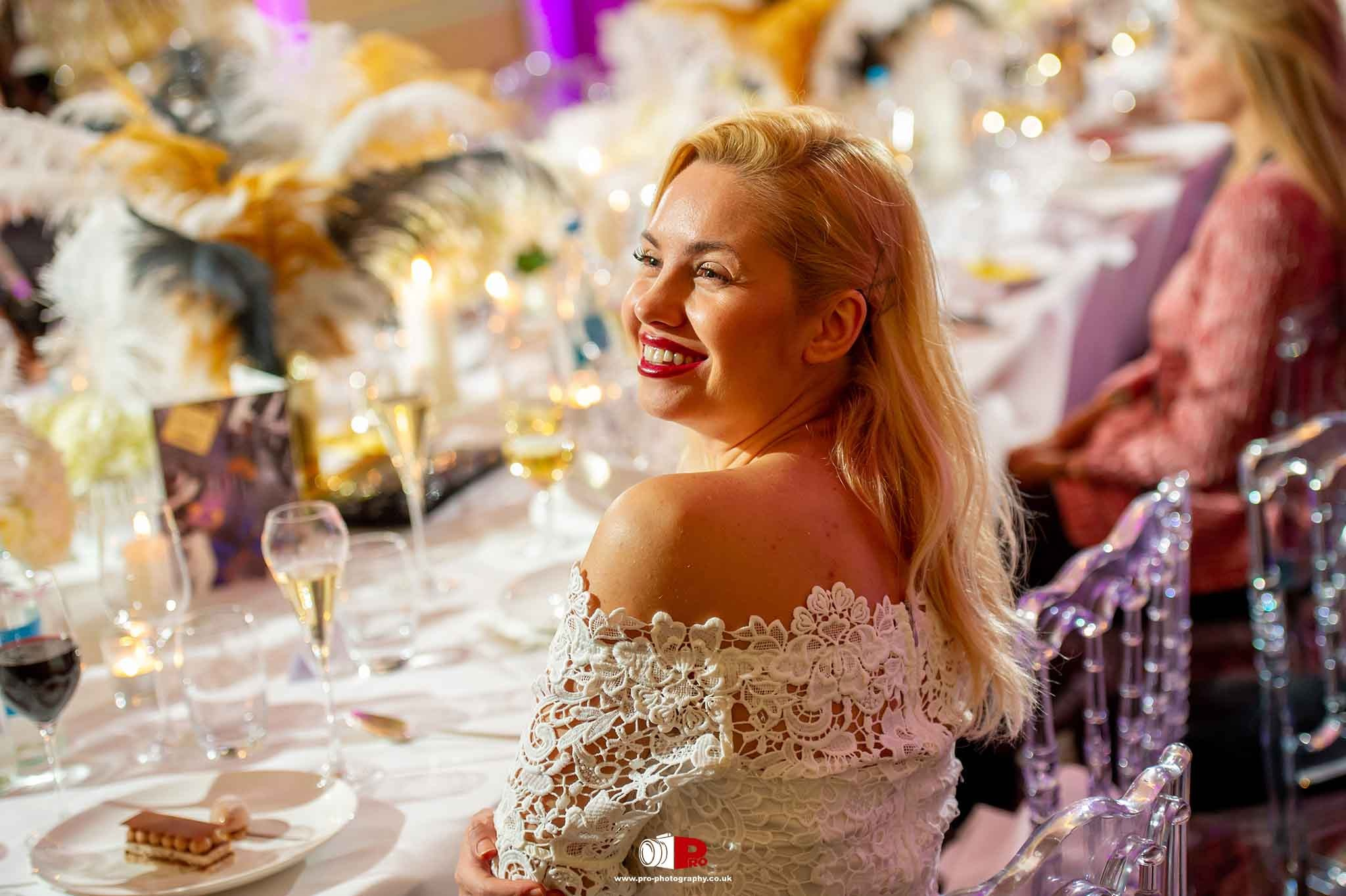  A smiling woman in a white lace dress enjoying a formal dinner with a beautifully decorated table in the background.