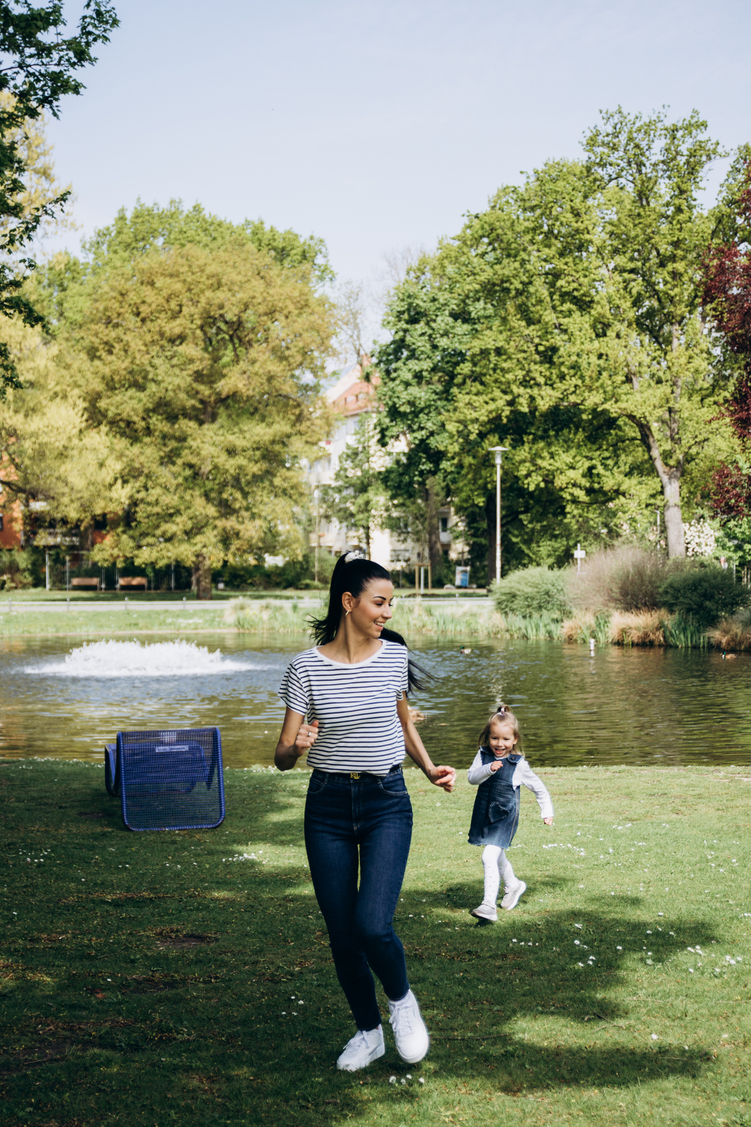Lebendige Momentaufnahme einer Mutter, die mit ihrer kleinen Tochter im Park spielt – natürliche Familienfoto Session bei Tageslicht am Wasser.