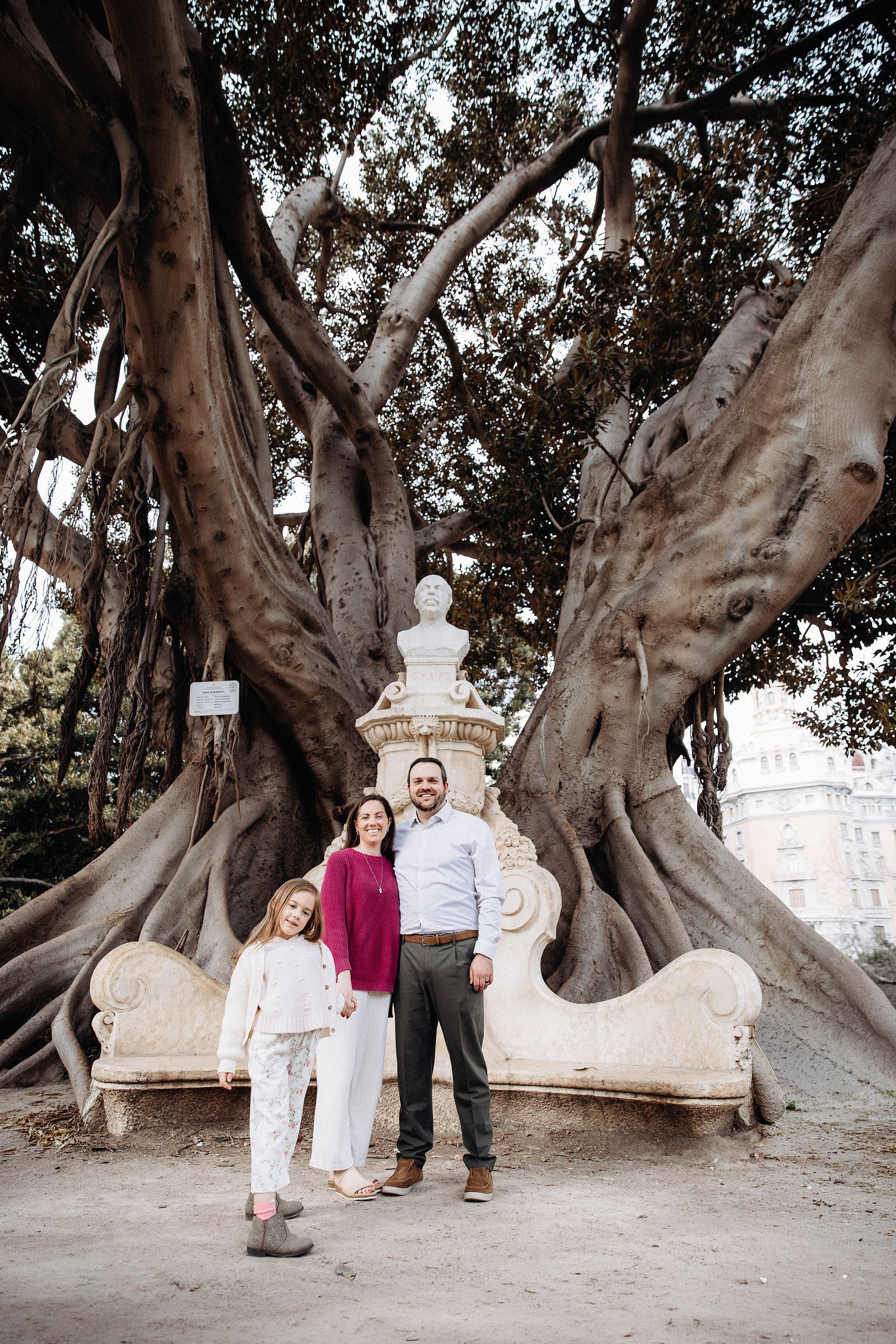 Семейная фотосессия в Валенсии, Испания, в зелёном историческом парке с большими скульптурными деревьями и декоративным фонтаном, где родители и ребёнок стоят вместе — естественная, живая семейная фотография в Валенсии, идеально подходящая для вневременных outdoor-портретов в Испании.