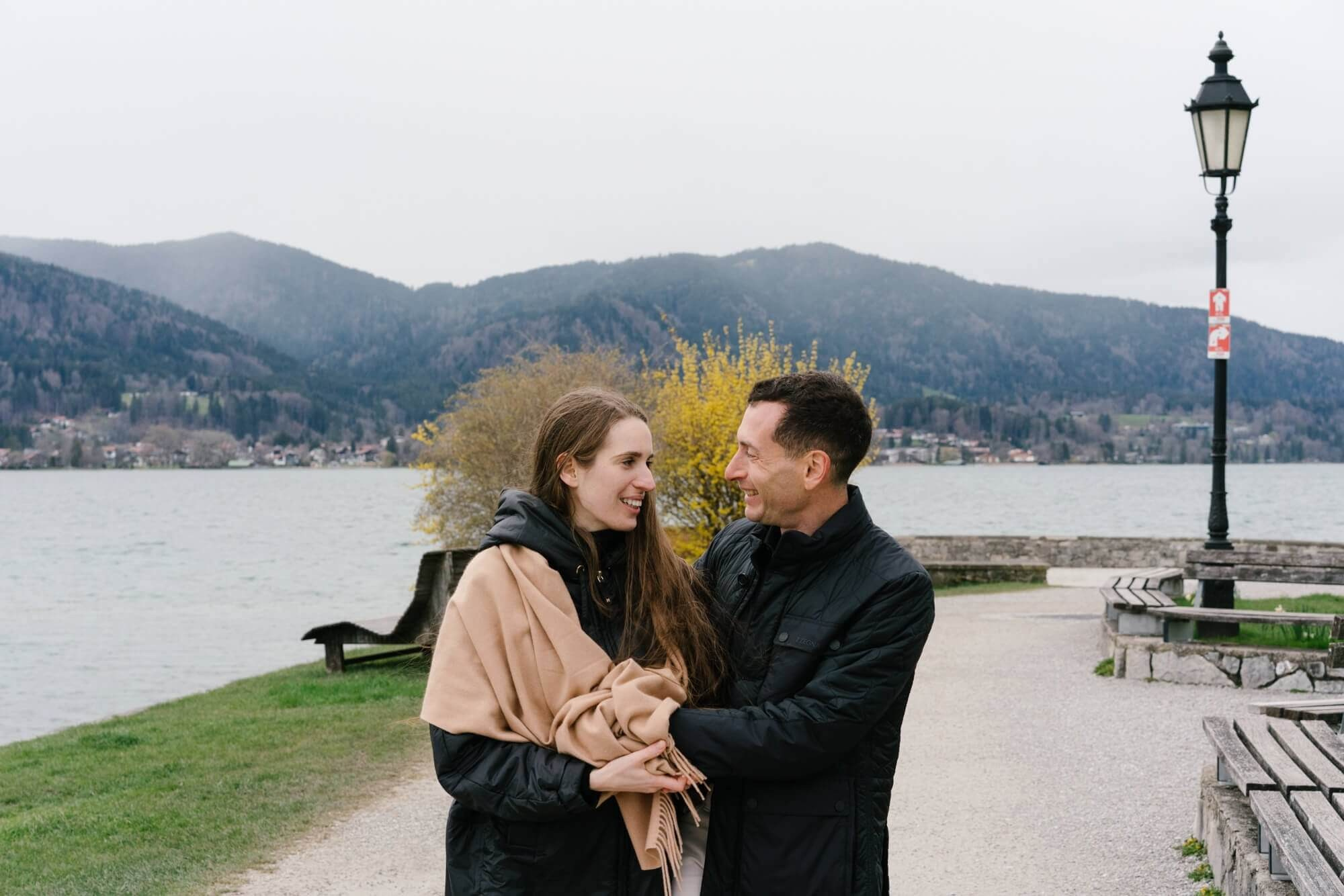 Couple standing close together at Tegernsee lake promenade with lamp post and mountains in background Germany