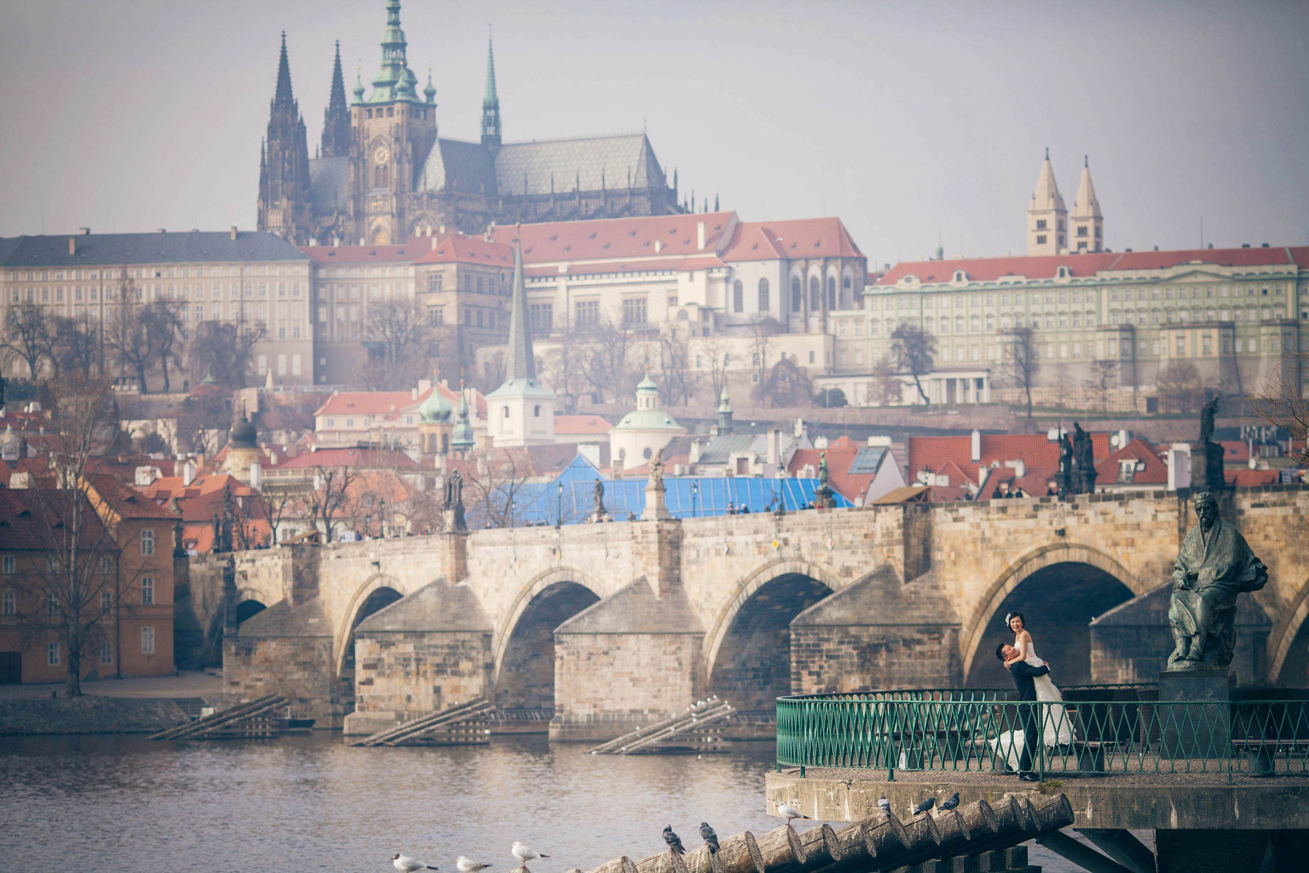 A groom wearing a tuxedo picks up his smiling bride at the riverside as near the Charles Bridge during their winter wedding celebration in Prague.