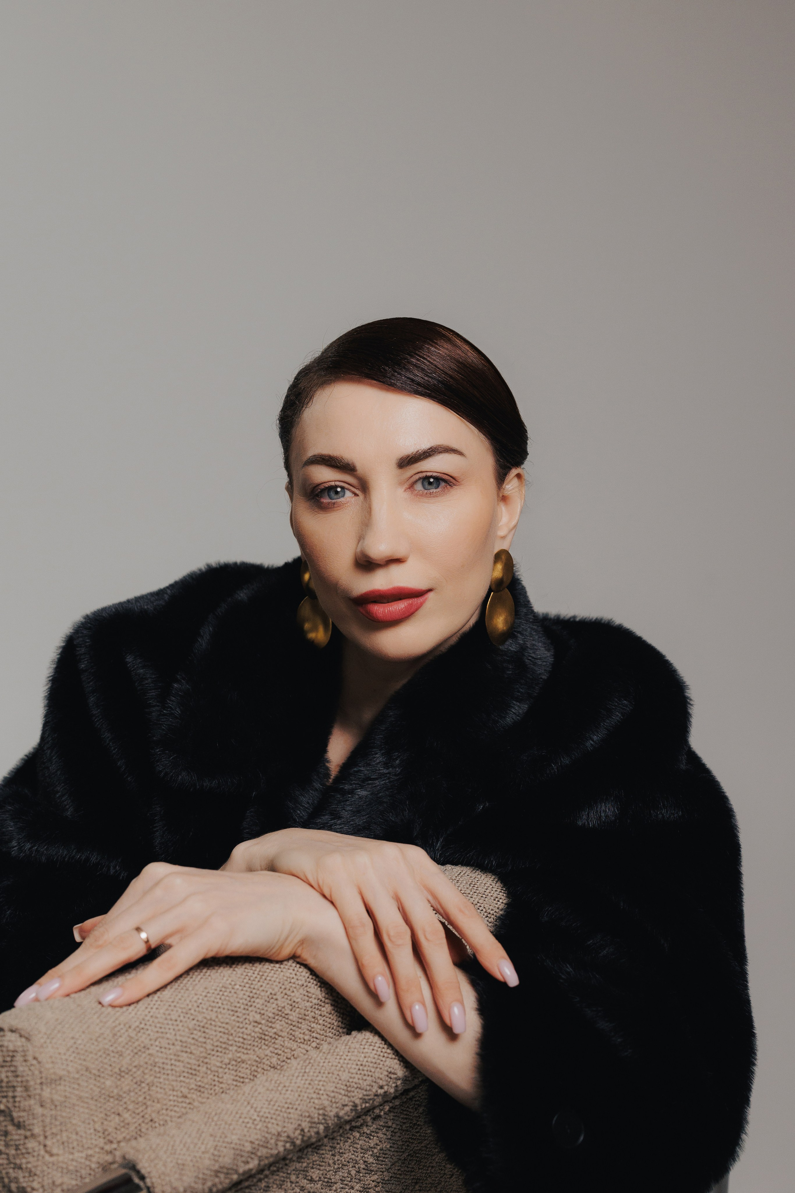 Elegant studio portrait of a woman in a black fur coat resting her arms on a chair with soft dramatic lighting, photographed in Leipzig, Germany. Элегантный студийный портрет девушки в чёрной шубе, опирающейся руками на кресло, в мягком драматичном освещении, снятый в Лейпциге, Германия.