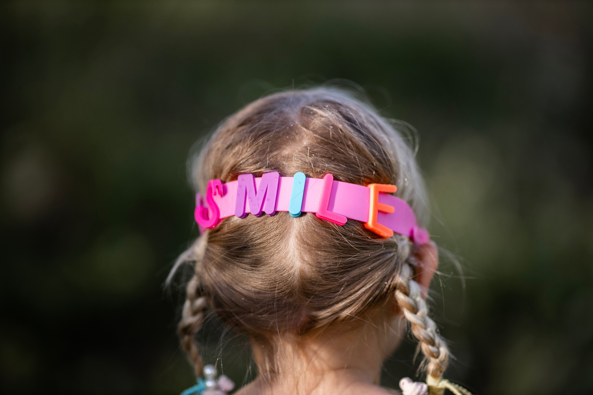 Zadnji deo glave malog deteta sa plavim kikama vezanim u repiće, koje nosi roze traku za kosu na kojoj šarenim slovima piše „SMILE”, na otvorenom.