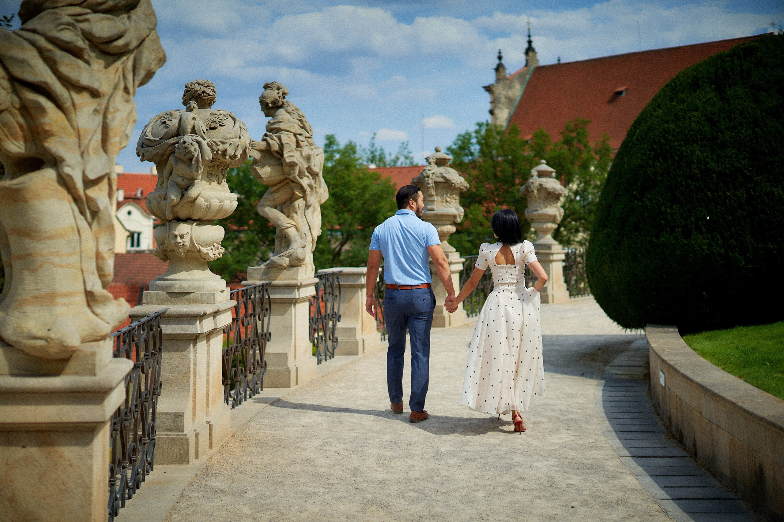 Couple walking hand-in-hand past classical statues as they exit Vrtba Garden, Prague.