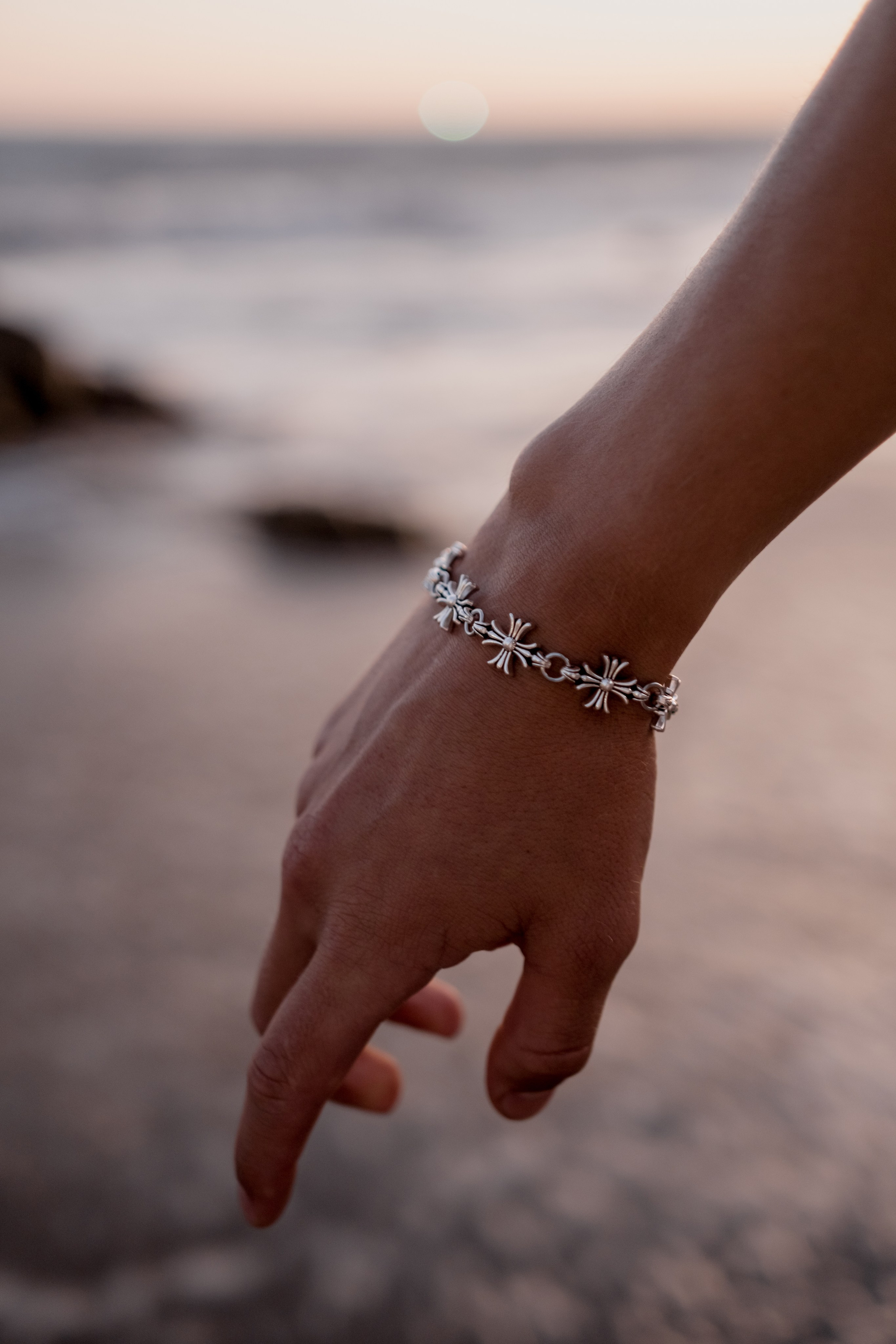 Close-up of a hand adorned with a silver bracelet reaching toward the ocean waves at sunset.