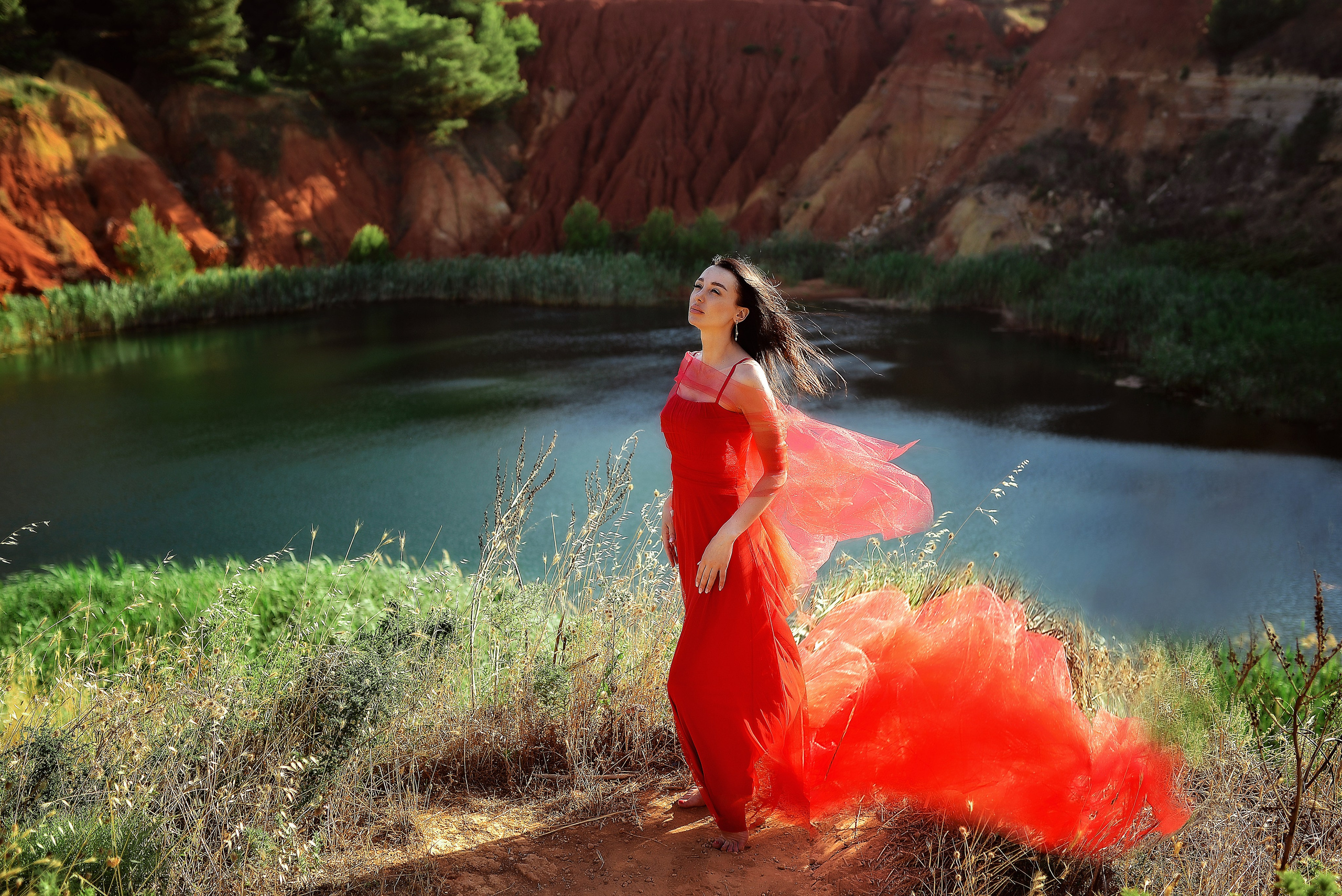 A woman in a flowing red dress stands on the edge of a scenic Bauxite Quarry, Otranto in Salento, her eyes closed and face turned toward the sun, with the vibrant train of her dress trailing in the breeze.