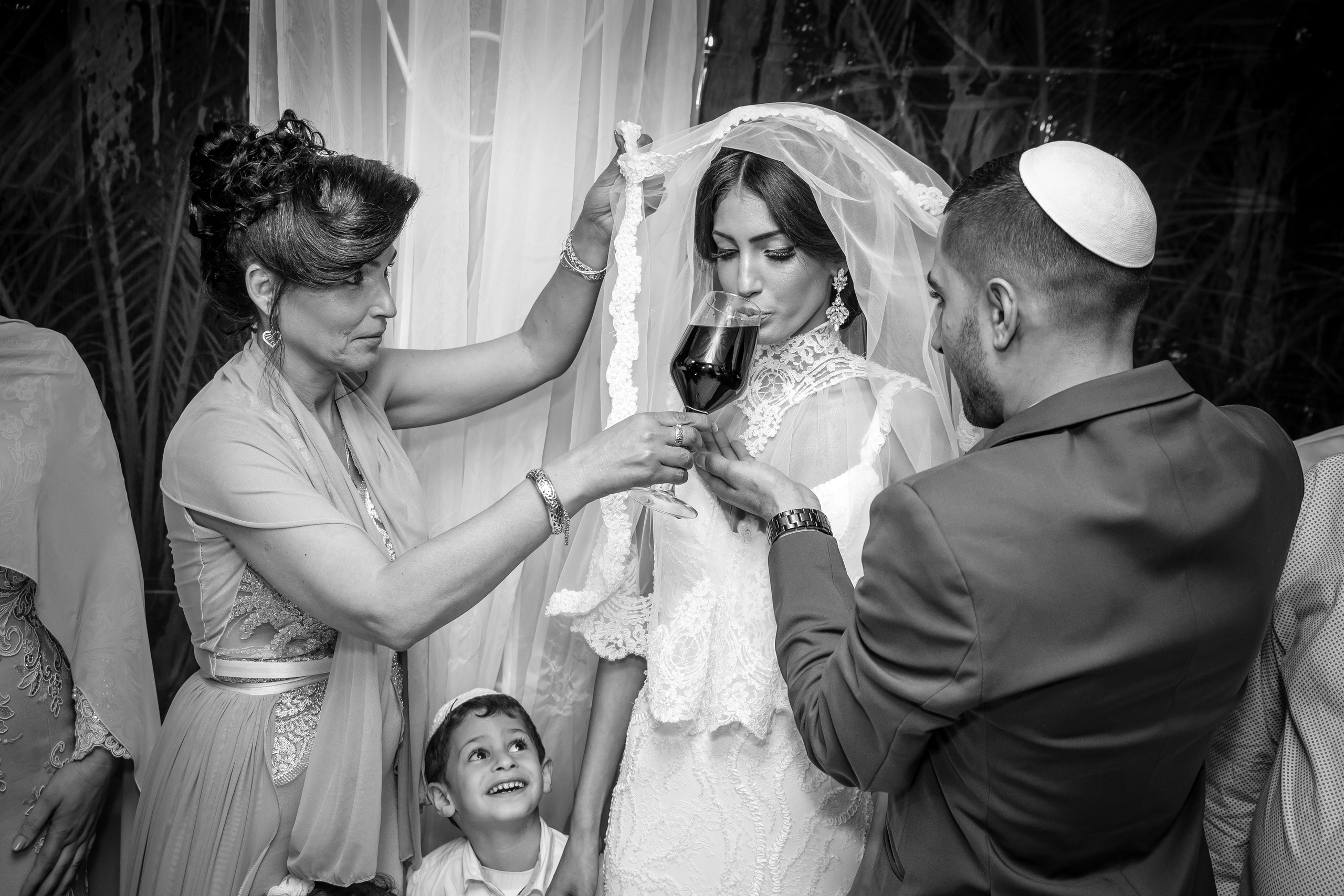 Jewish wedding ceremony: the bride drinks wine as the groom and a woman assist, with a smiling boy in the foreground