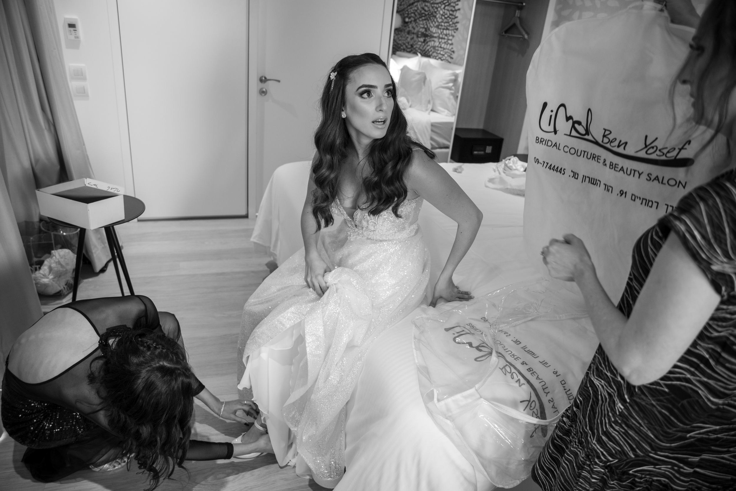 Bride sitting on the bed while a bridesmaid helps her with shoes, surrounded by loved ones in a candid wedding preparation moment.