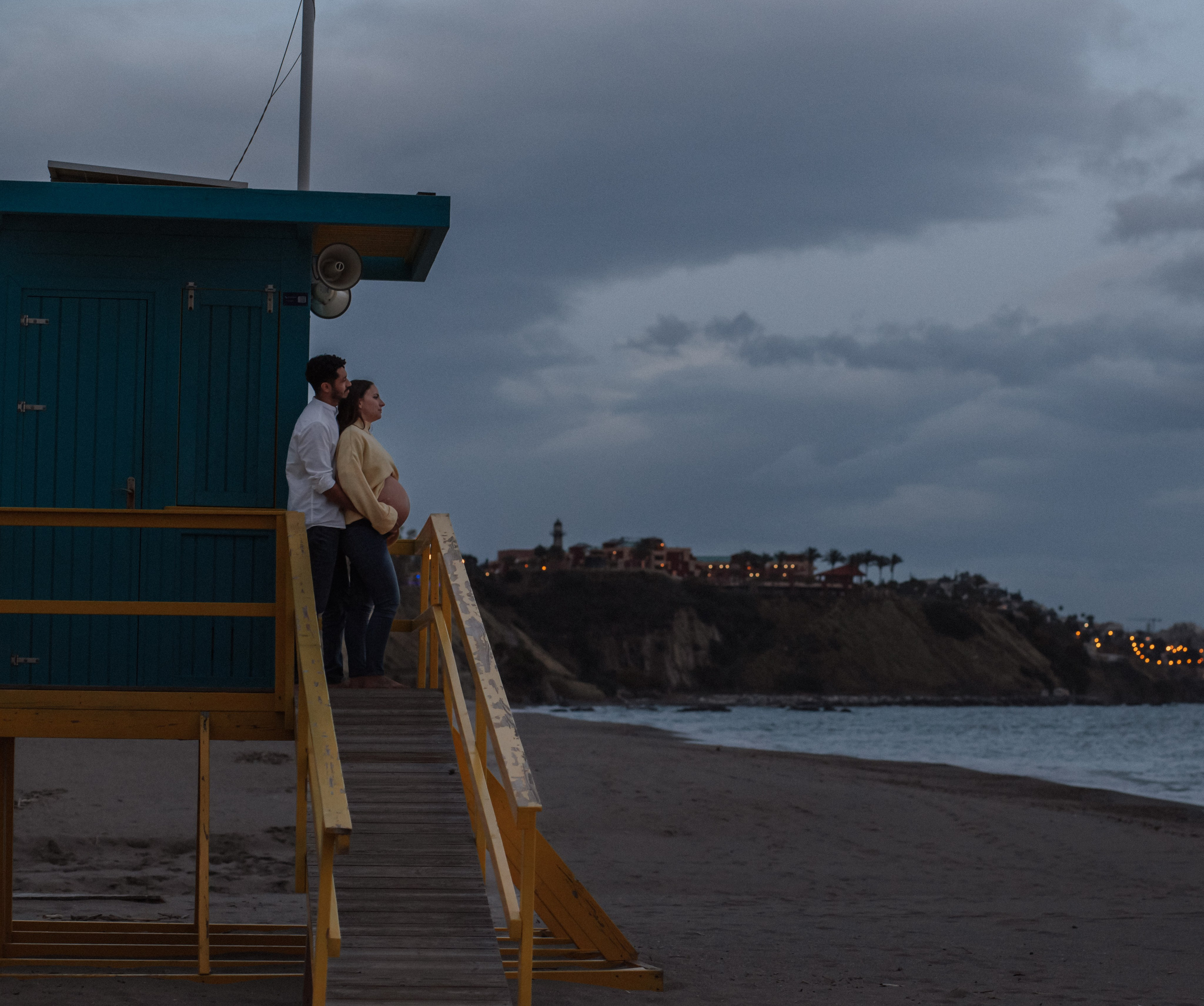 Sesión romántica de embarazo en Málaga, España — pareja embarazada de pie en una torre de socorrista al anochecer, con el faro y la costa de fondo. Perfecta para quienes buscan fotografía creativa de embarazo en Málaga y la Costa del Sol.