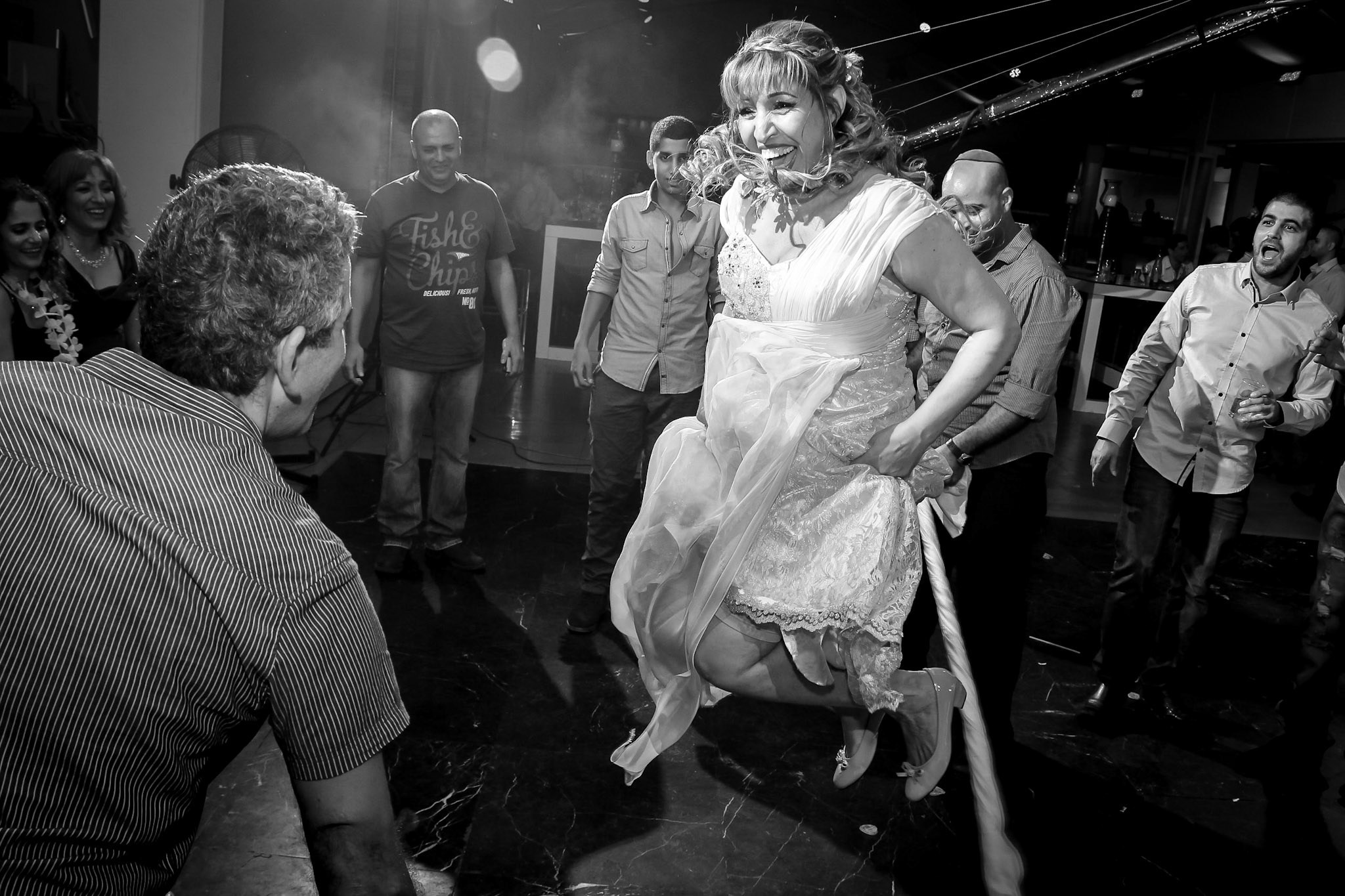 A joyful mother of the bride mid-air as she jumps over a rope during a wedding celebration, surrounded by cheering guests. The black-and-white photograph captures the energy, movement, and excitement of the moment.